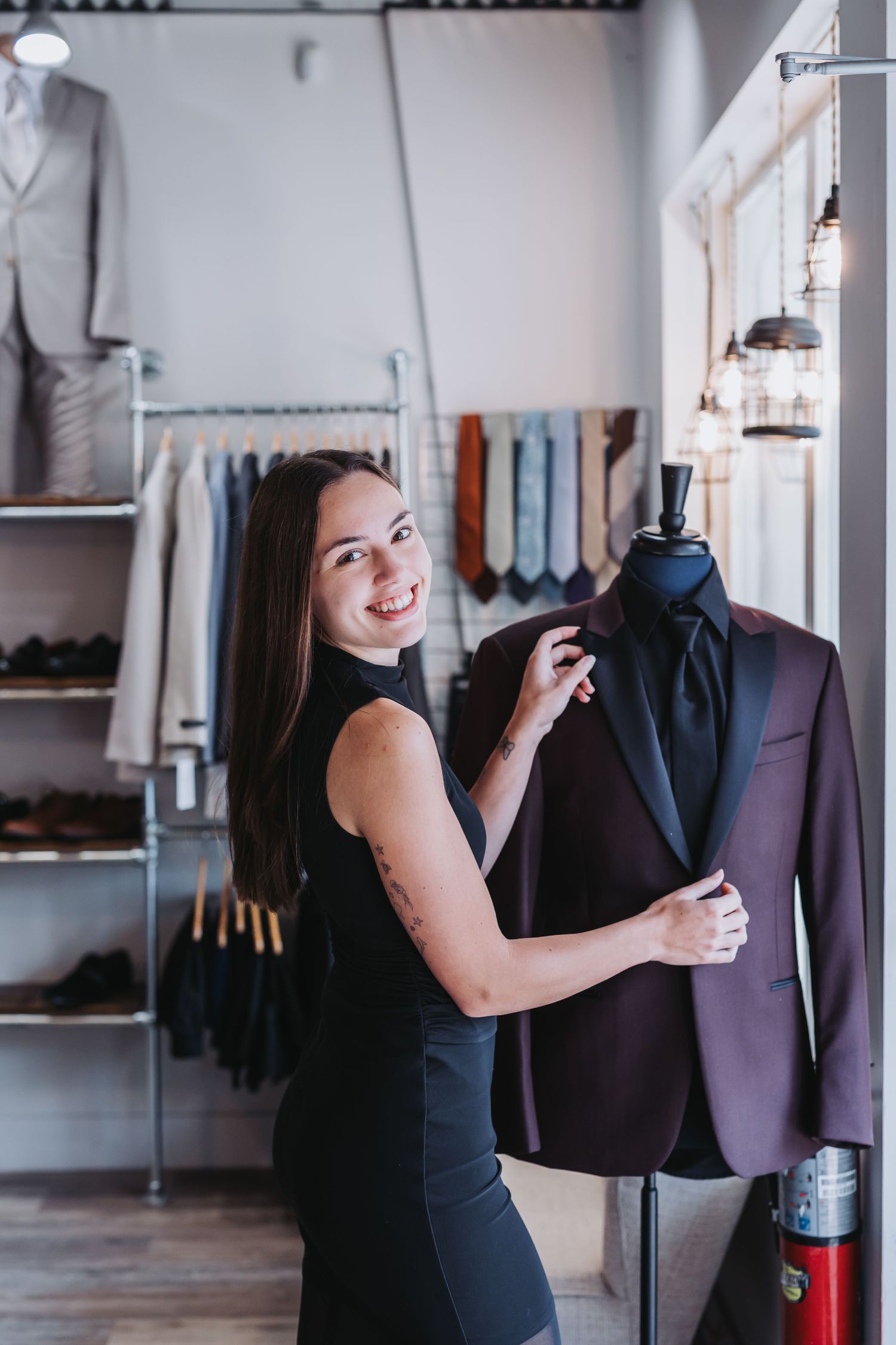 Smiling woman in black dress adjusting a burgundy tuxedo jacket on a mannequin in a modern menswear boutique.