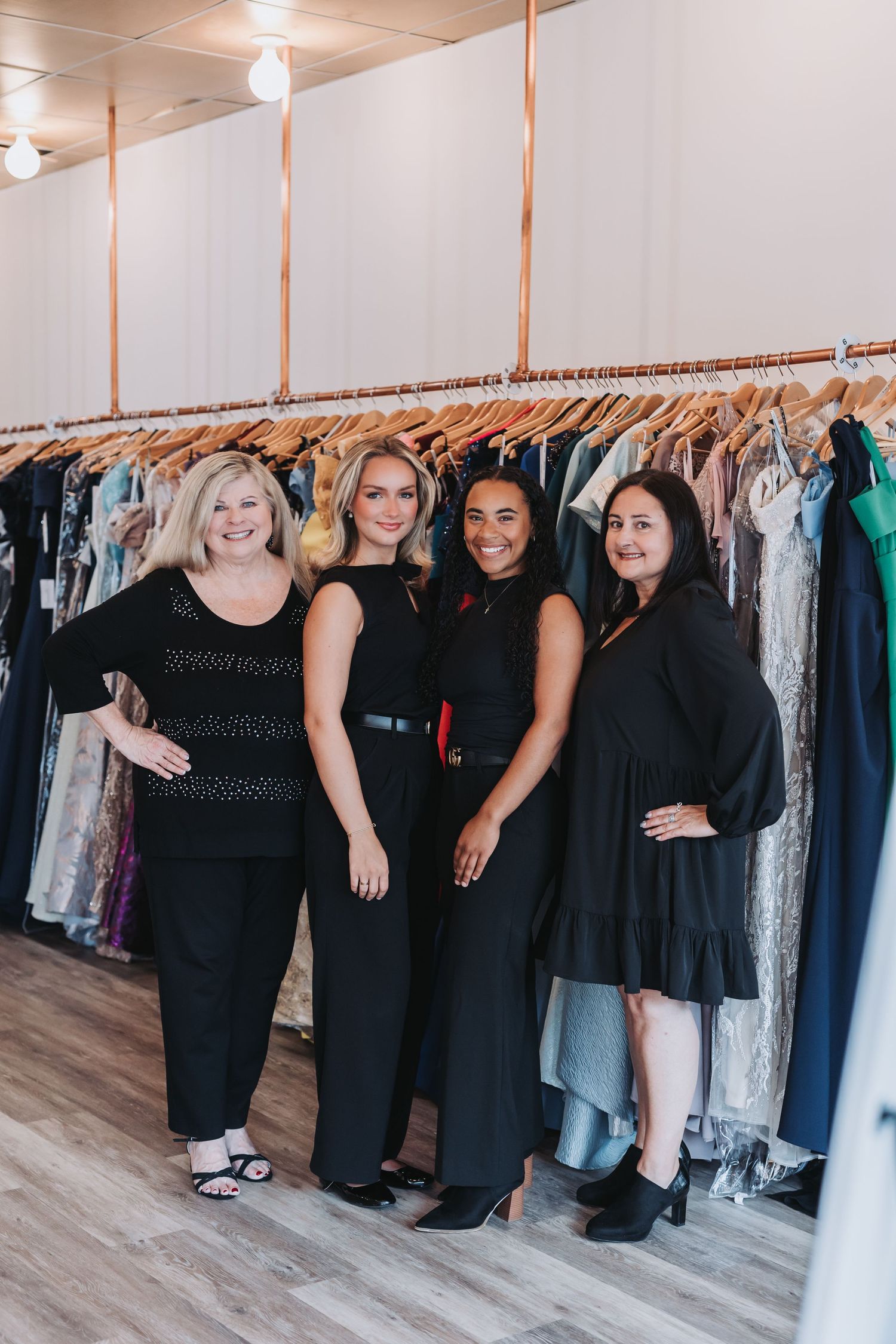 Four women in black outfits smiling in a boutique dress shop with colorful gowns on racks behind them.