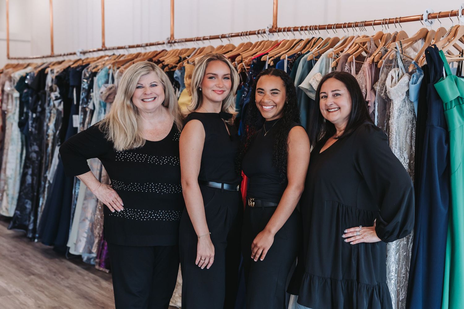 Four women in black outfits smiling together in a boutique clothing store with dresses on racks behind them.