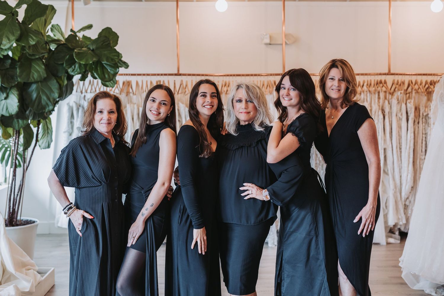Six women in matching black outfits pose together smiling in an elegant bridal boutique with wedding gowns displayed behind them.