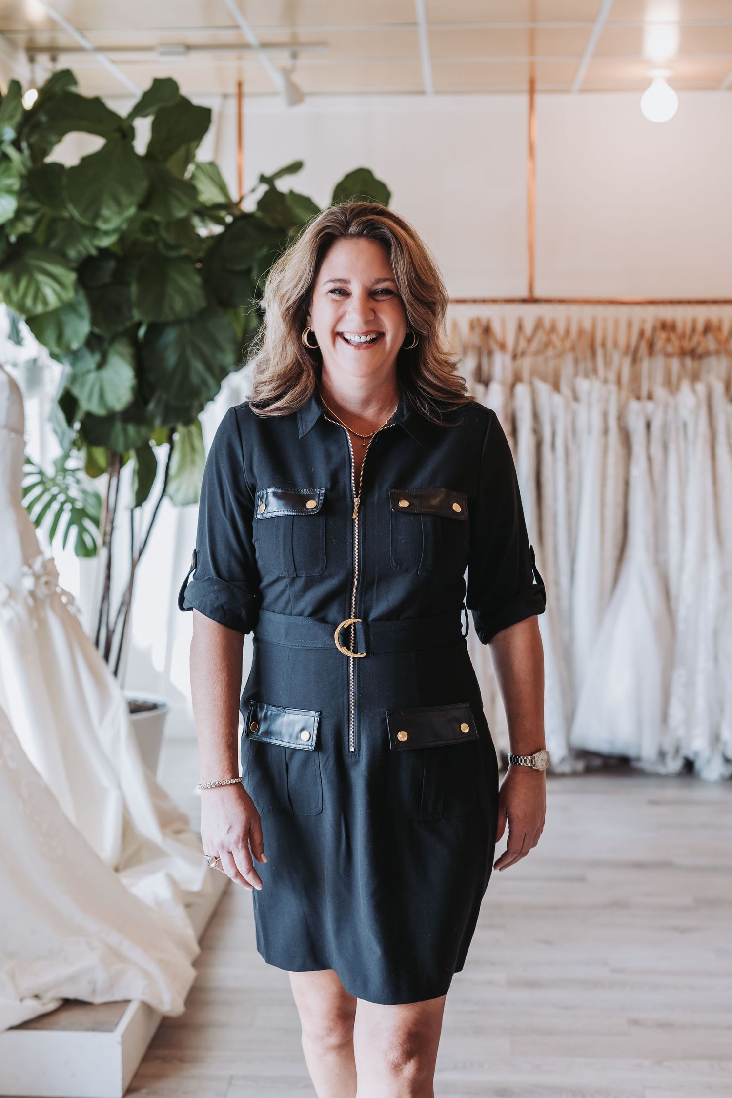Smiling woman in black belted dress standing in a bridal boutique with wedding gowns on racks in background.