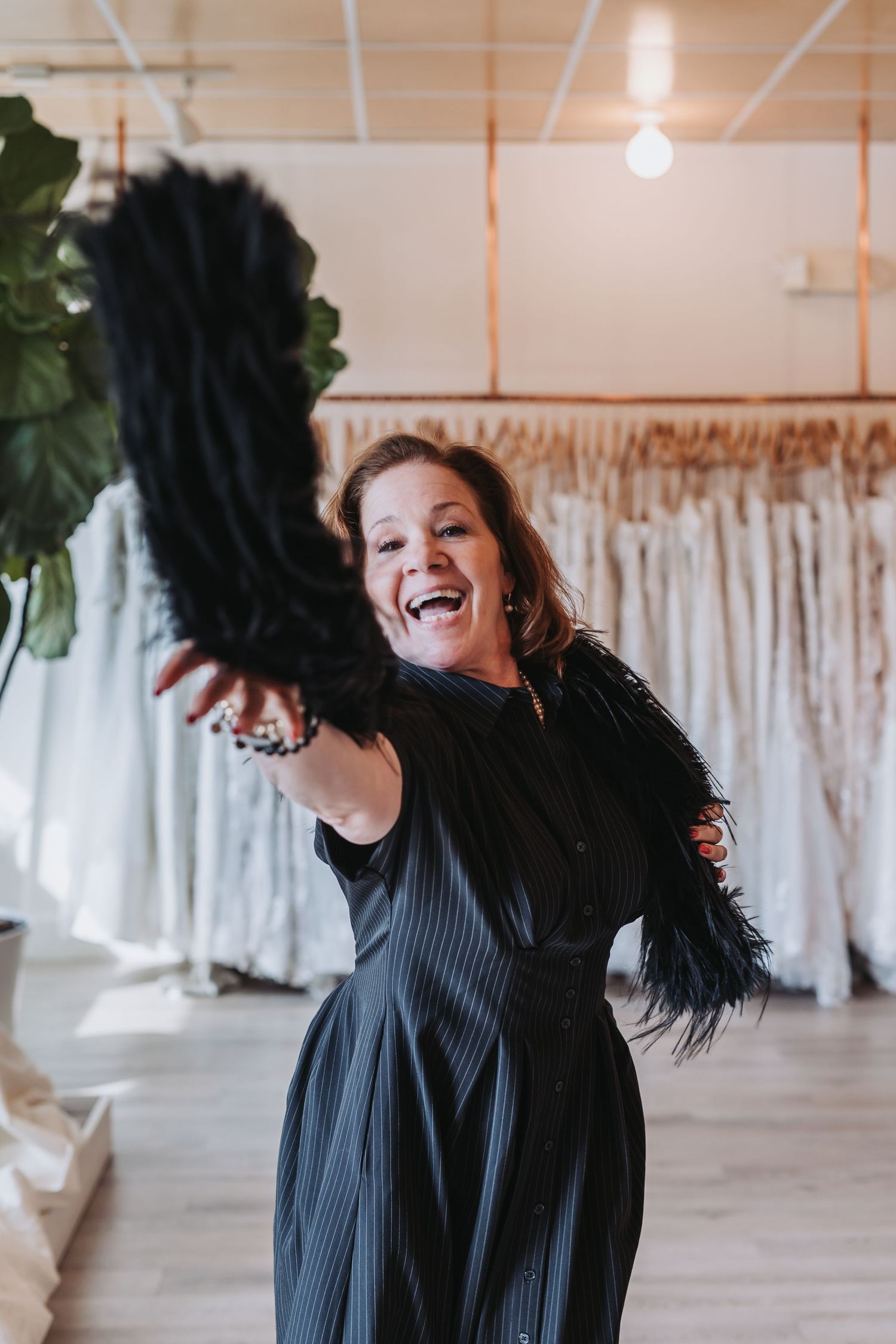 A joyful woman in a black feathered dress laughs while holding a feather fan in a bridal boutique.