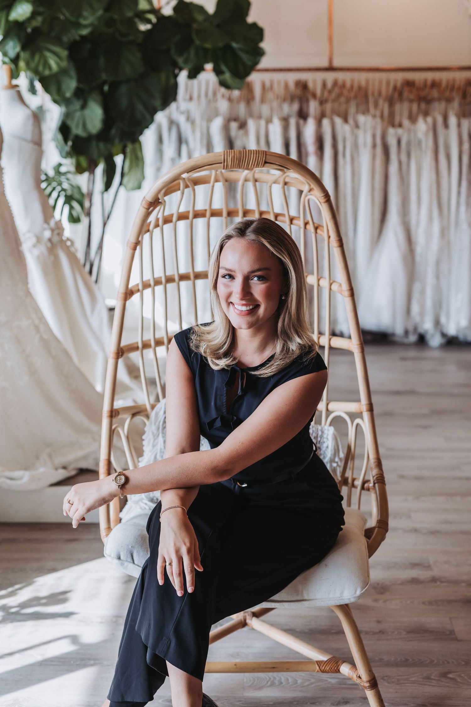 Smiling blonde woman sitting in rattan chair inside a bridal boutique with wedding dresses displayed in background.