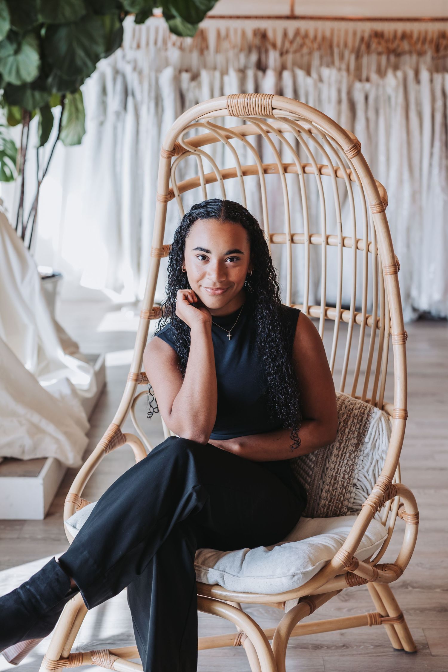 Woman in black outfit sitting in rattan egg chair inside a bridal boutique with wedding dresses in background.