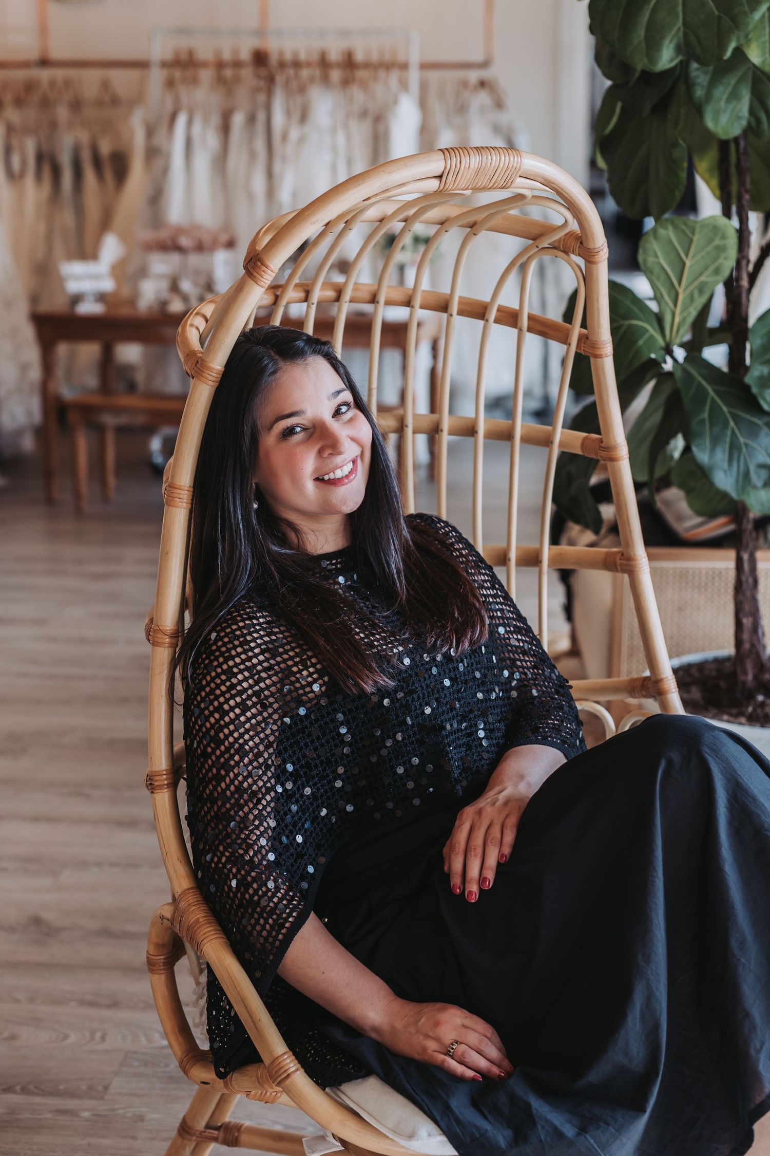Smiling woman in black lace top sitting in rattan egg chair inside a bright boutique shop.