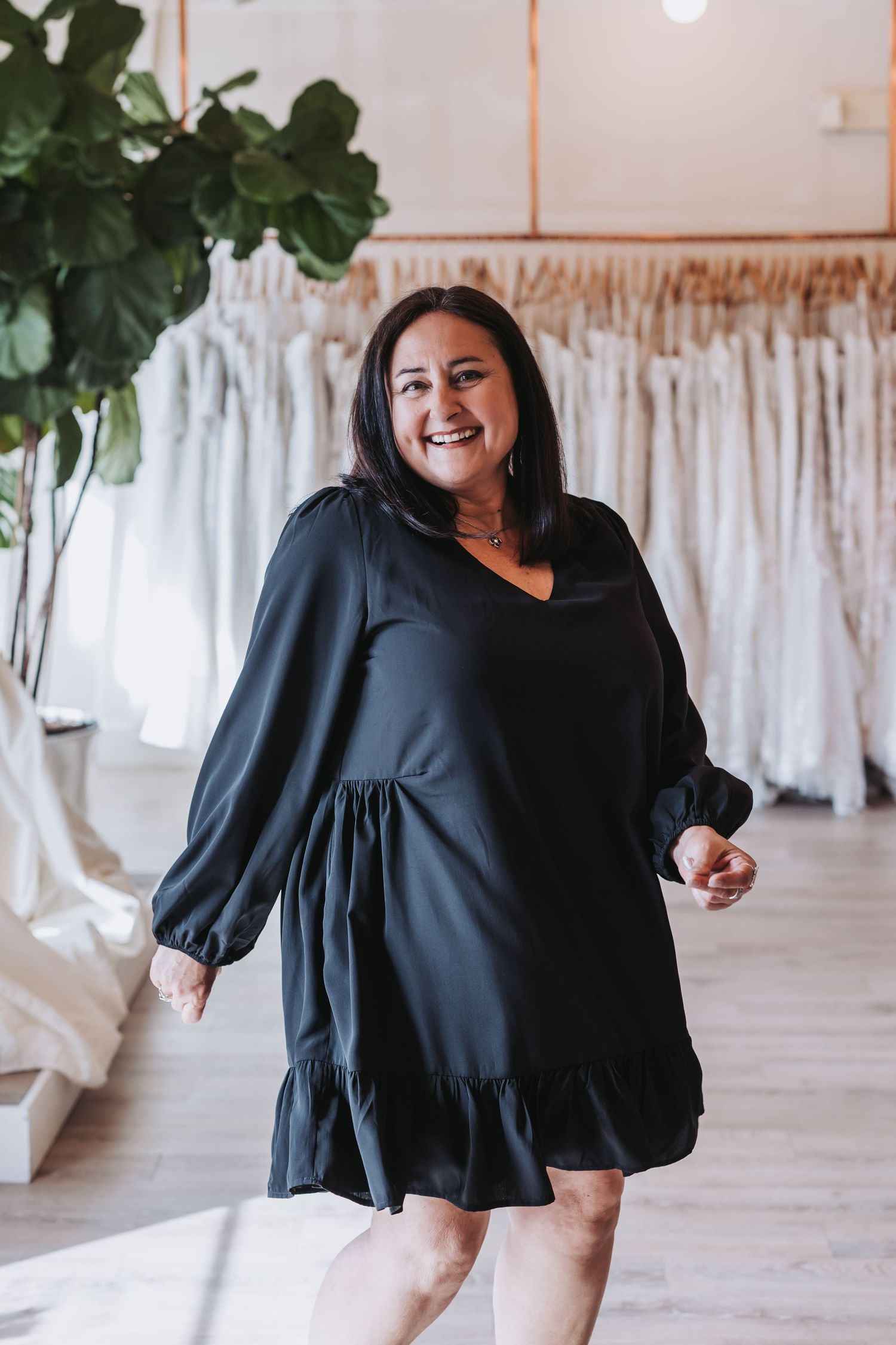 Woman in black long-sleeve mini dress smiling in a bridal boutique with wedding gowns displayed in the background.