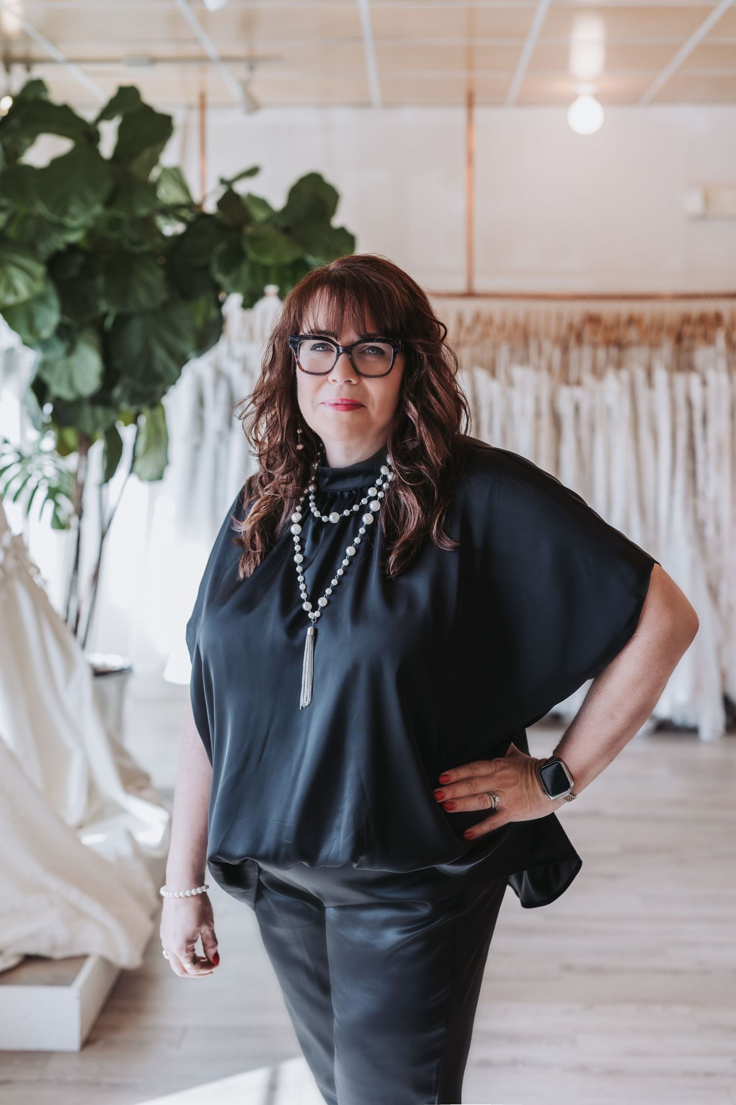 Woman in black top and leather pants poses confidently in a bridal boutique surrounded by wedding gowns.