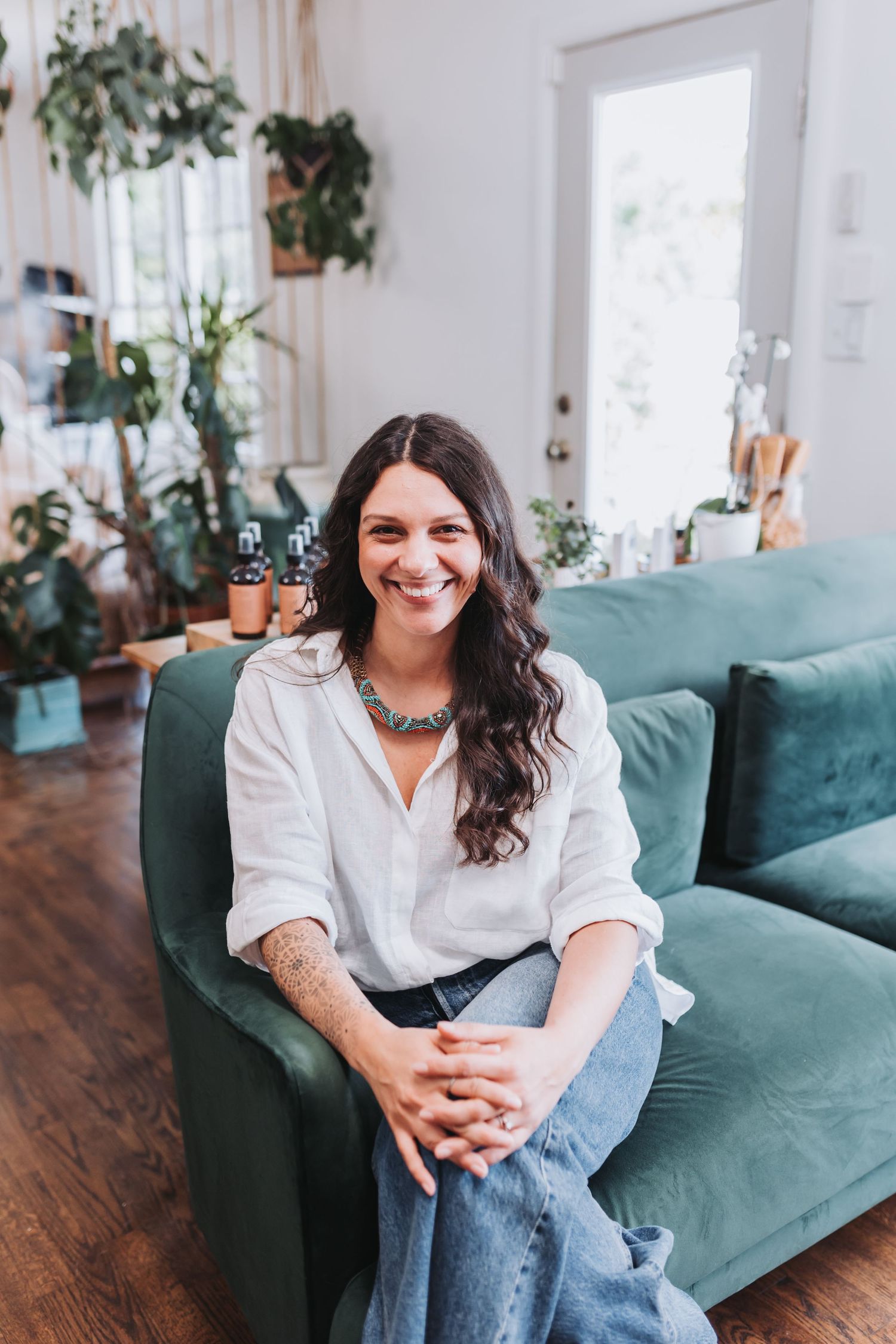 Smiling woman with dark hair sits on a teal velvet sofa in a plant-filled living room, wearing a white shirt and jeans.