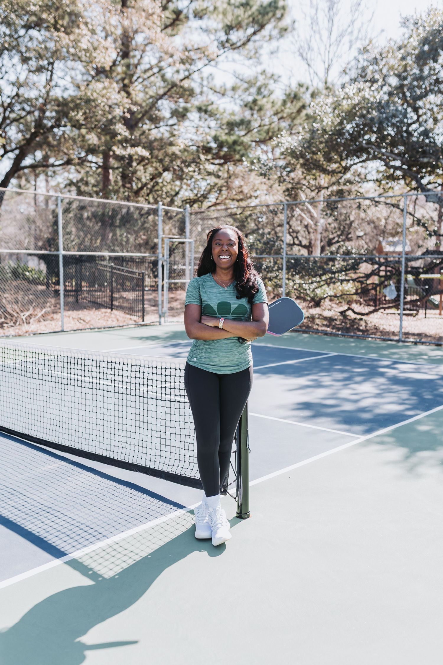 Woman smiling on pickleball court holding paddle, wearing teal shirt and black leggings near the net.
