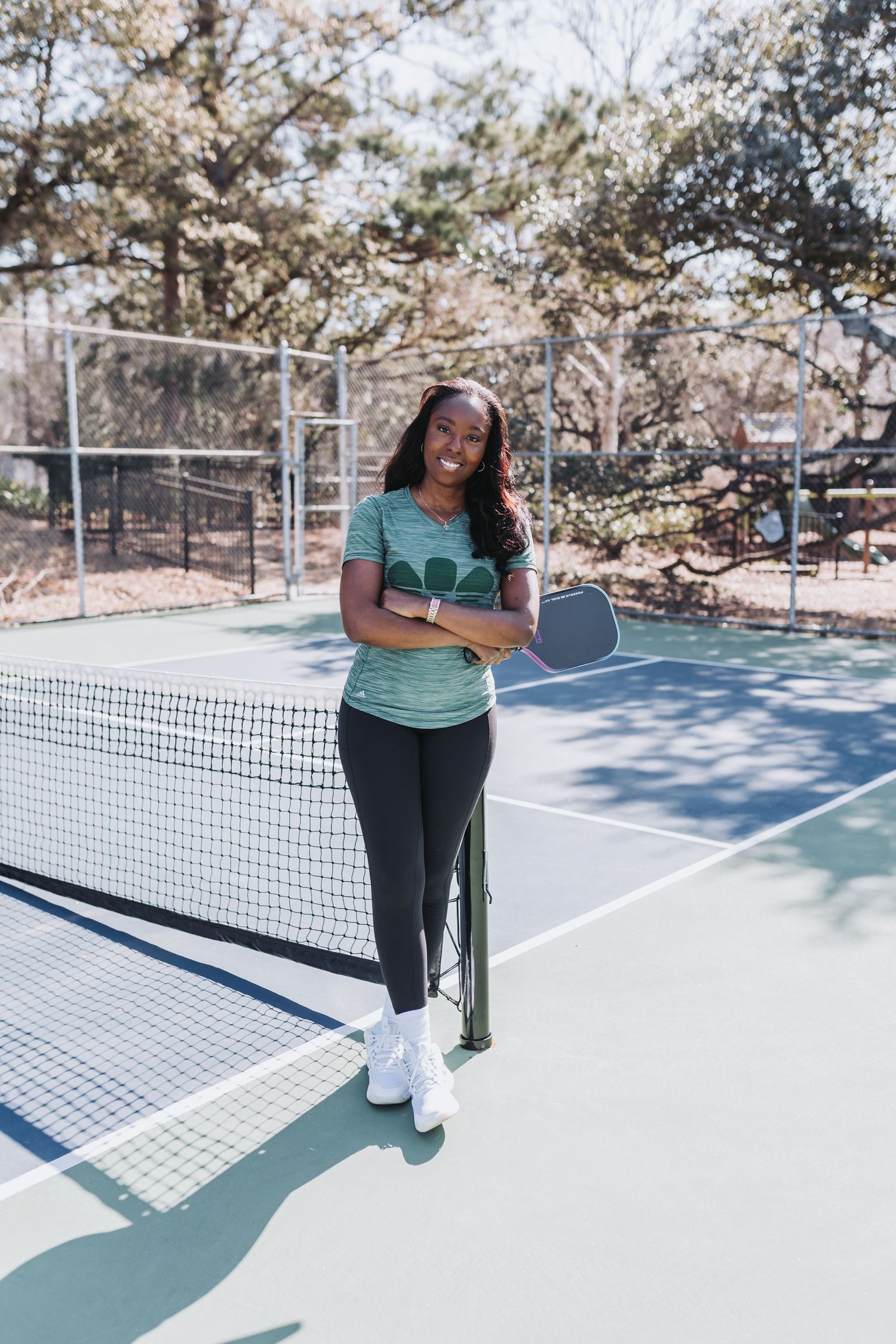 A smiling woman holds a pickleball paddle while standing at the net on an outdoor pickleball court.