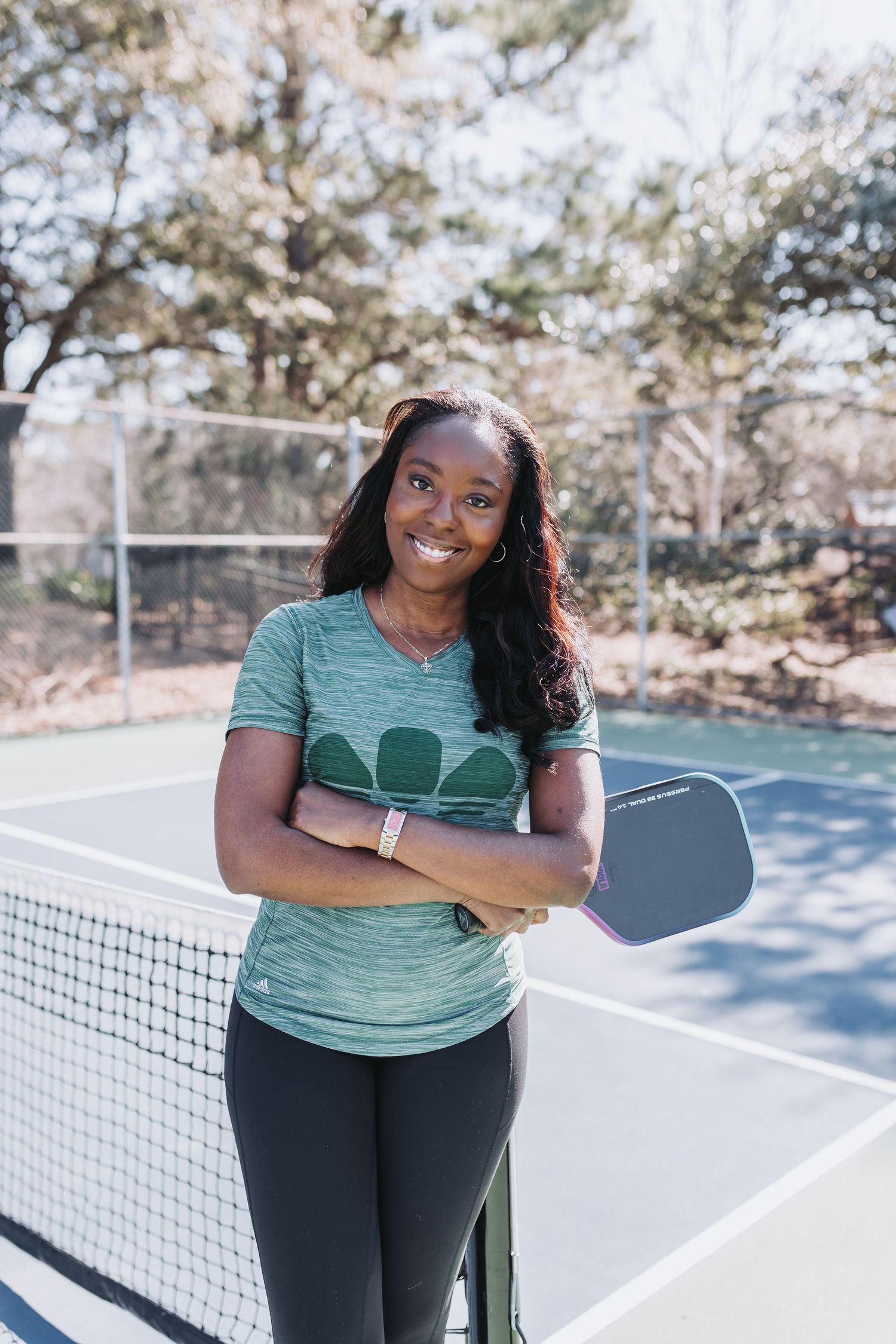 Smiling woman holding a pickleball paddle with arms crossed at an outdoor court.