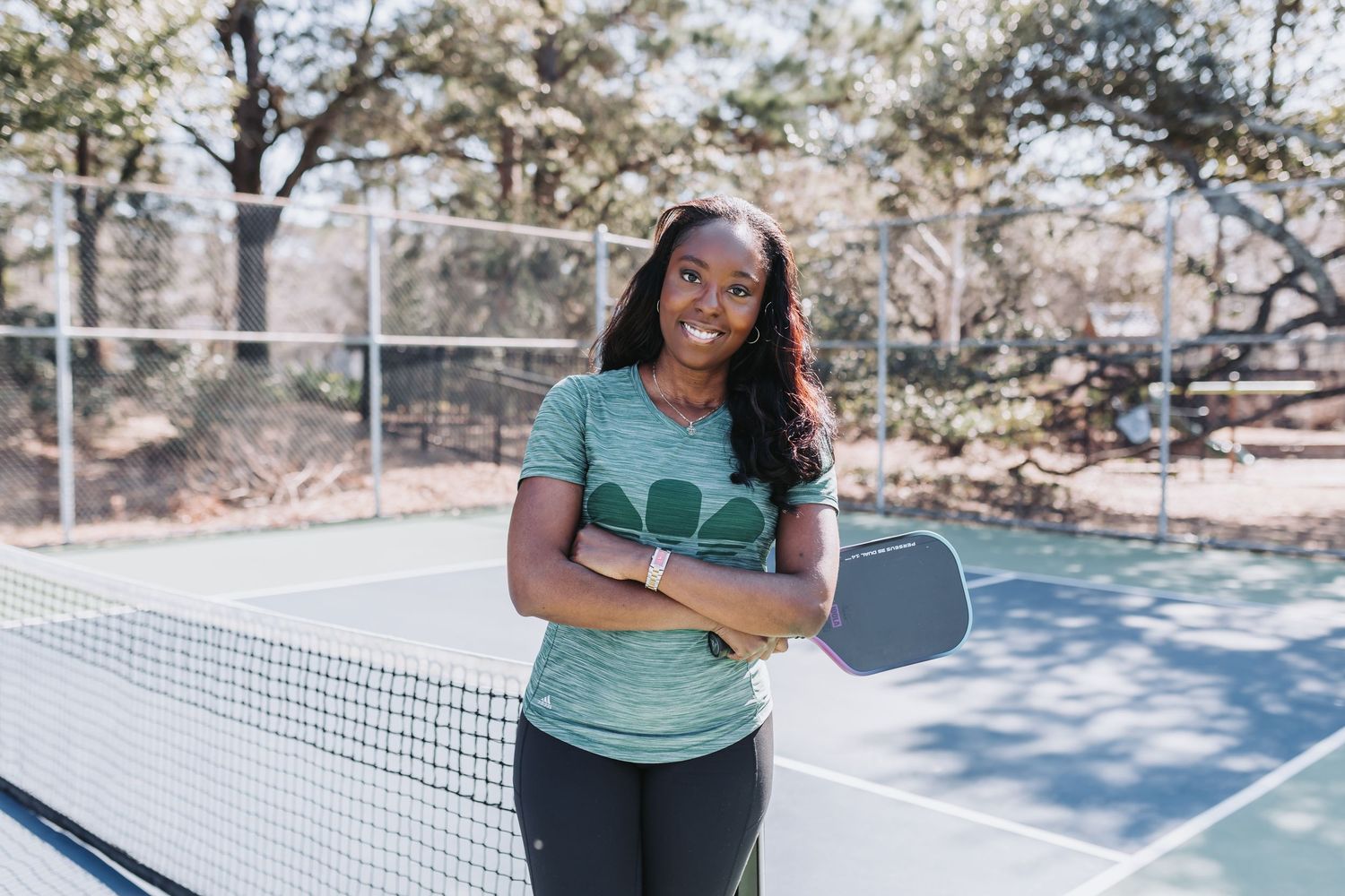 Woman smiling on pickleball court holding paddle, wearing green athletic shirt and dark leggings outdoors.