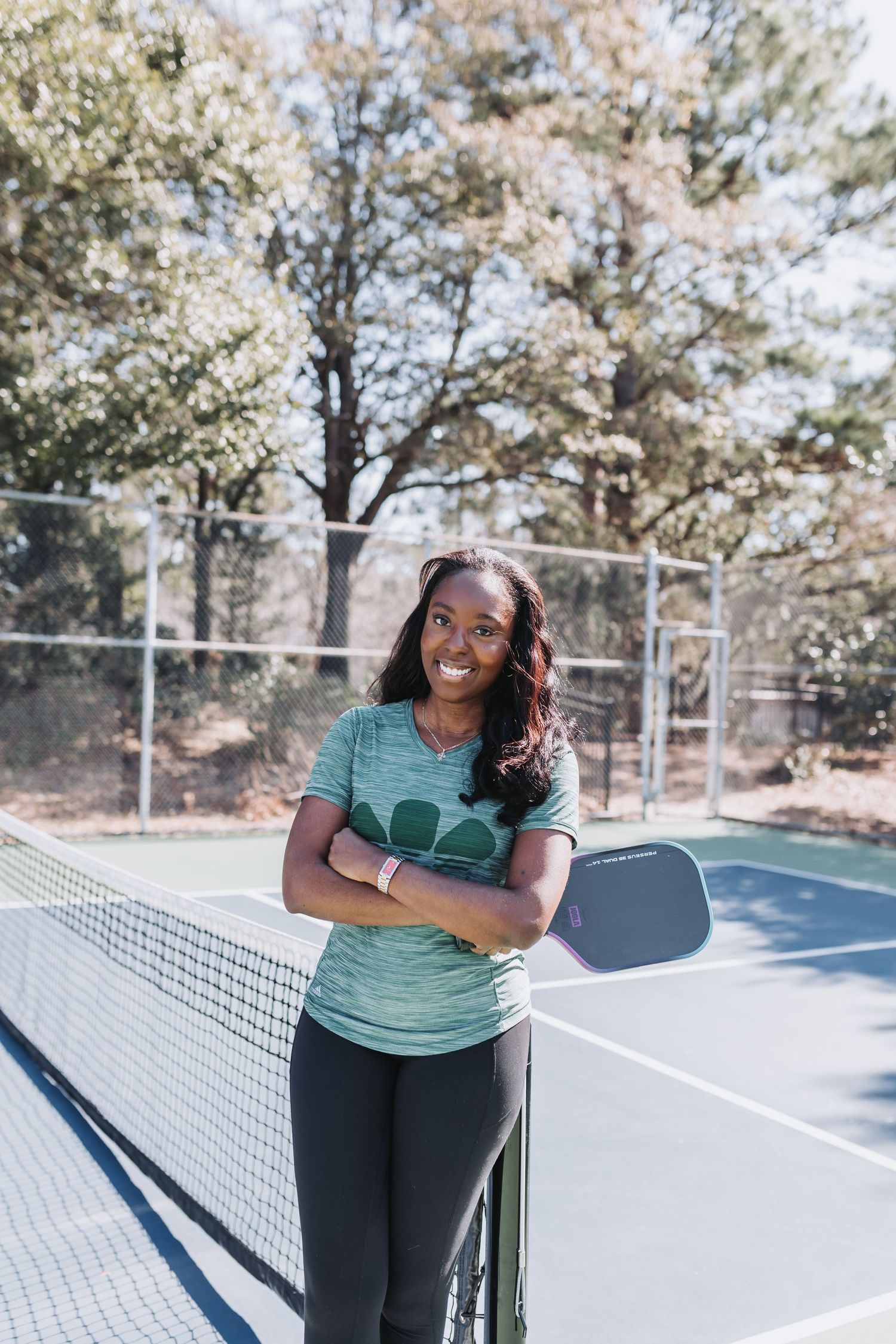 Happy woman posing with a pickleball paddle at an outdoor blue court surrounded by trees.