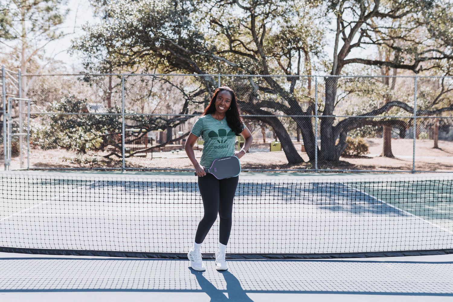 Woman in green t-shirt and black leggings posing confidently at a tennis court net outdoors.