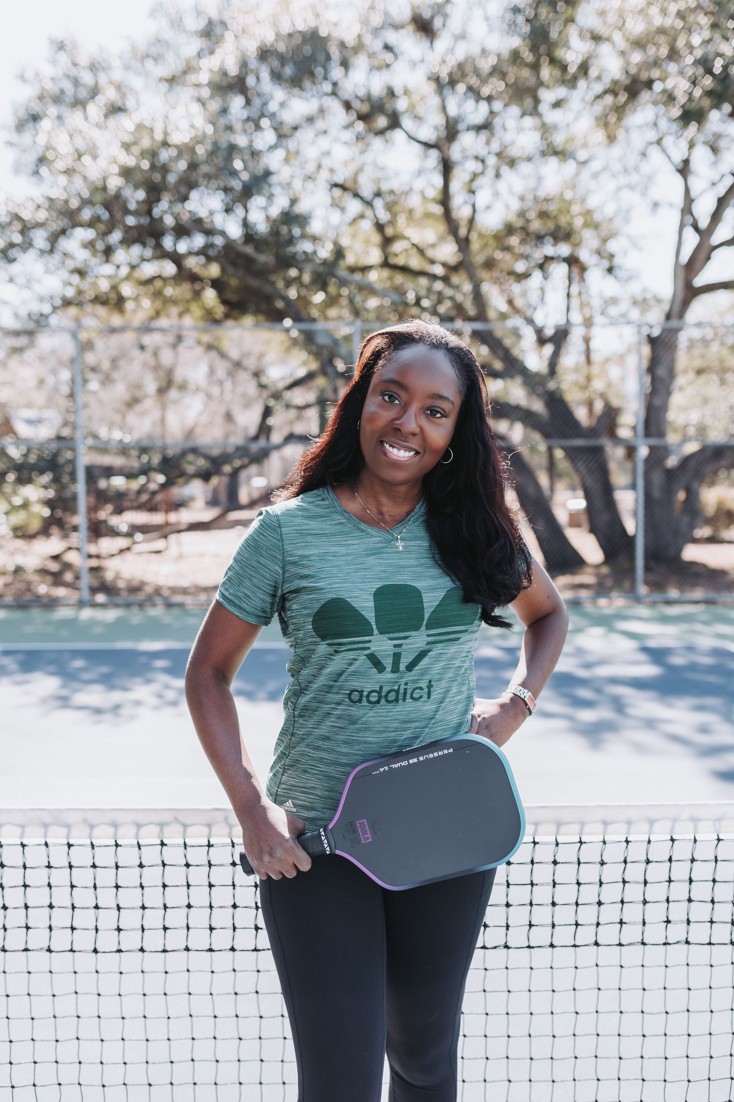 Smiling woman holding a pickleball paddle on an outdoor court, wearing a green Adidas-style pickleball t-shirt.