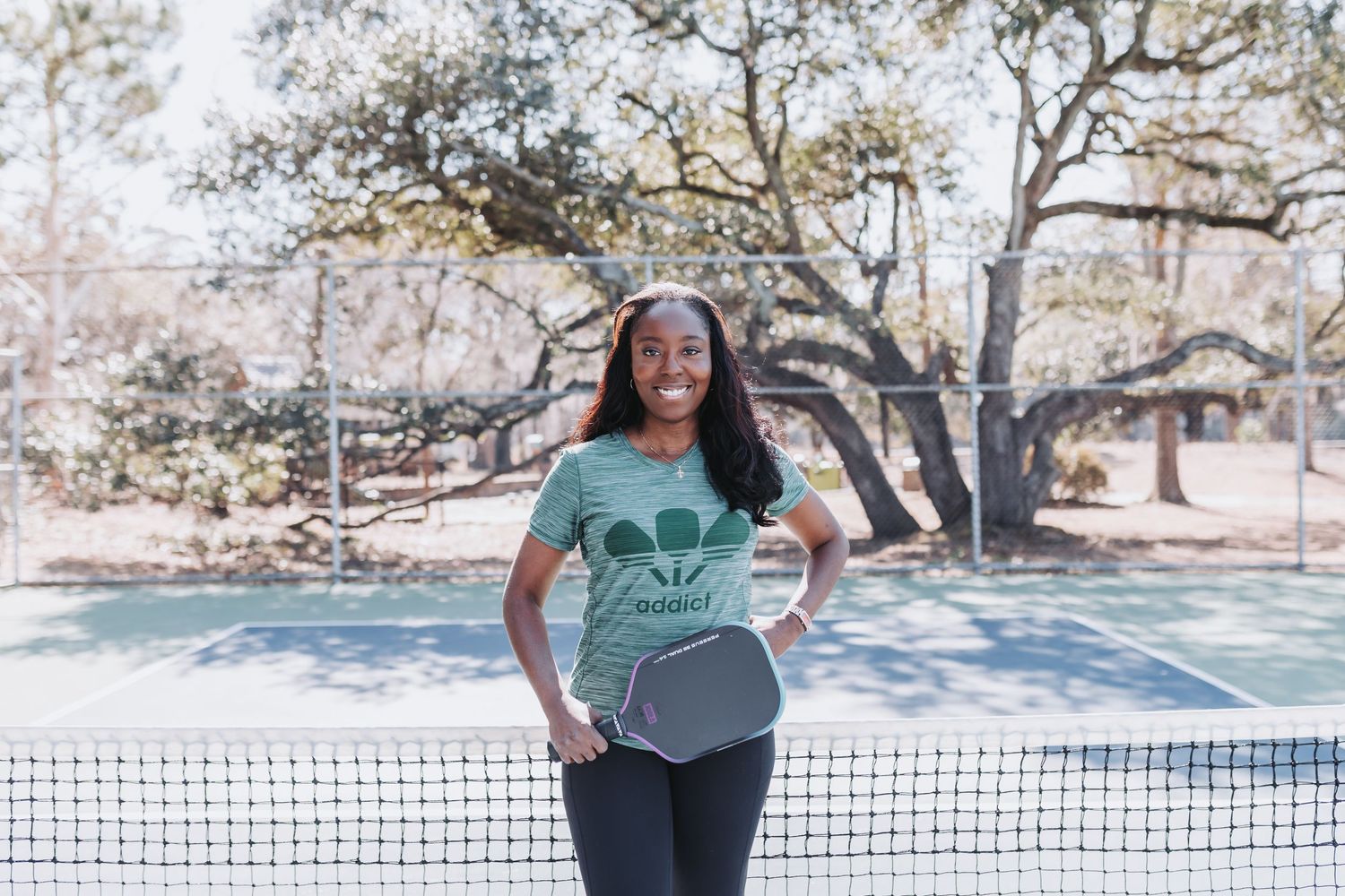 Woman smiling on pickleball court holding paddle, wearing green tee and leggings, trees in background.