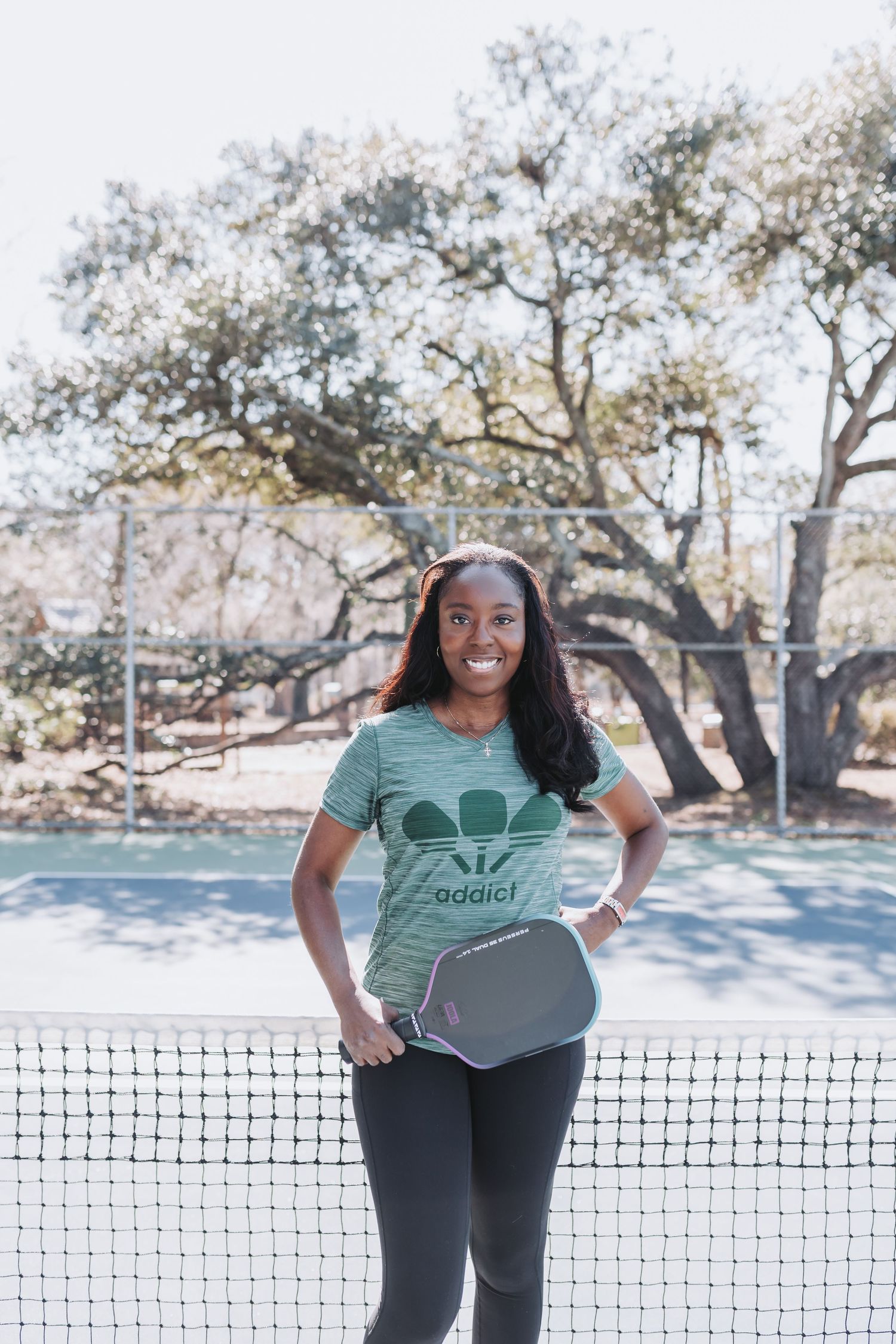 Woman in green Adidas shirt holding a pickleball paddle, standing at a court net outdoors.