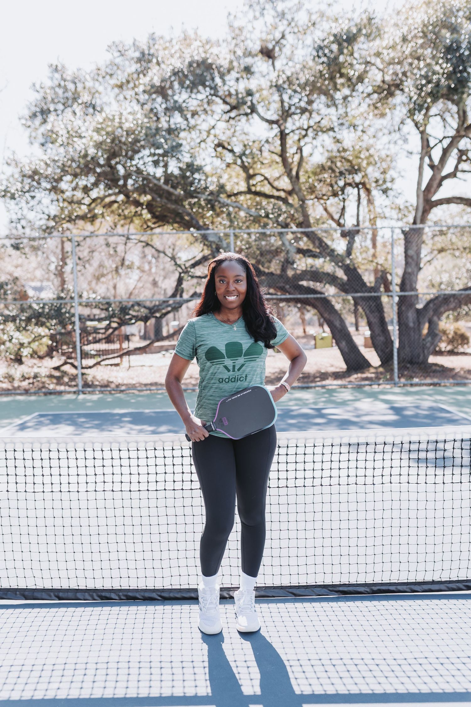 Smiling woman holding a pickleball paddle on an outdoor court, wearing a green graphic tee and black leggings.