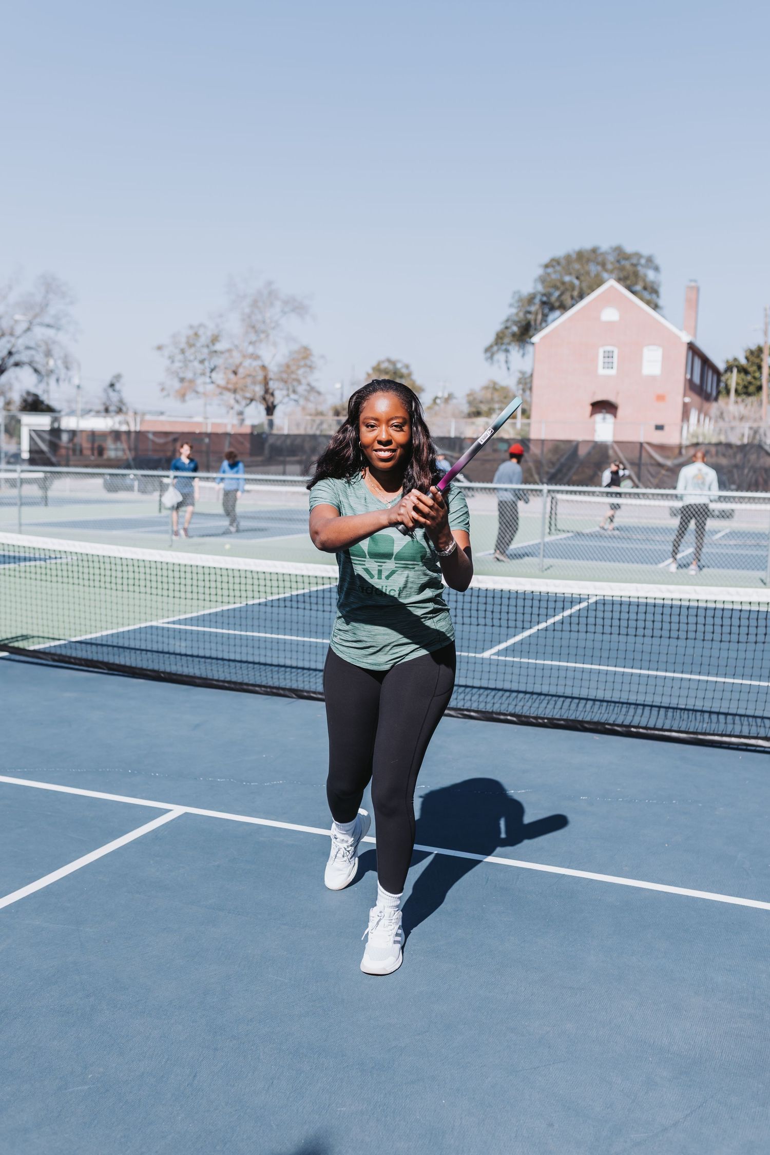 Young woman smiling on a blue tennis court, holding a racket, wearing a green shirt and black leggings.