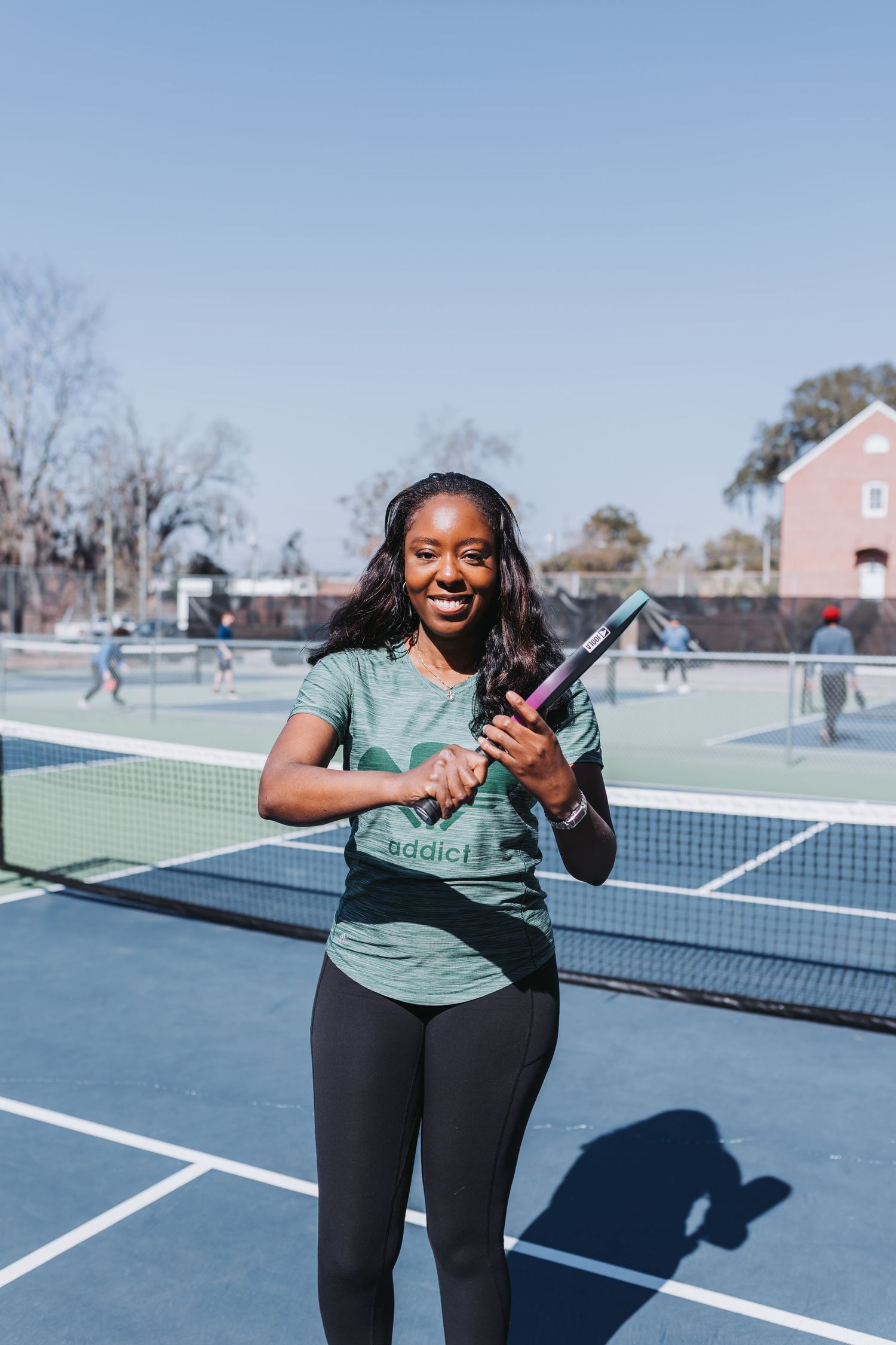 Smiling woman holding a pickleball paddle on an outdoor court wearing a green addict shirt and black leggings on a sunny day.