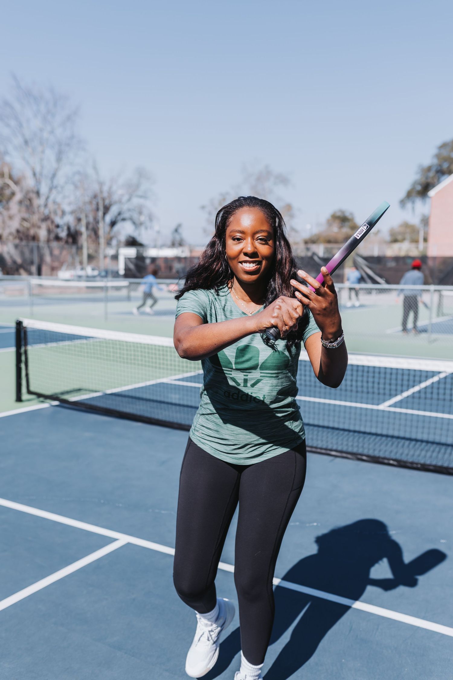 Smiling woman in green shirt holding tennis racket on blue outdoor court on a sunny day.