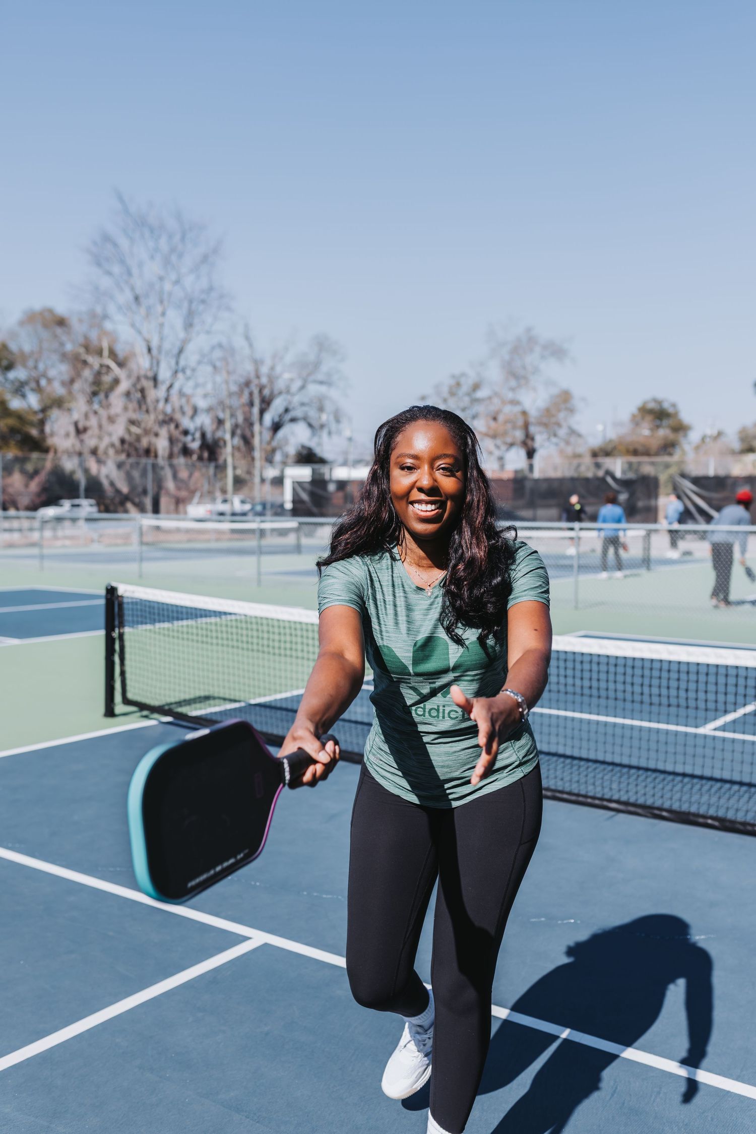 Smiling woman playing pickleball on an outdoor court, holding a teal paddle while wearing a green shirt and black leggings.