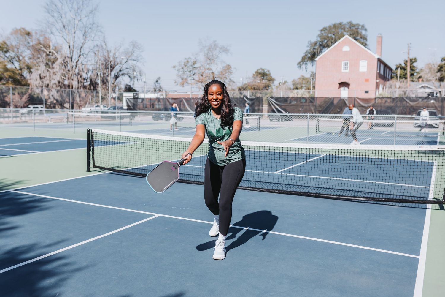 Woman playing pickleball on an outdoor blue court, smiling and holding a paddle on a sunny day.