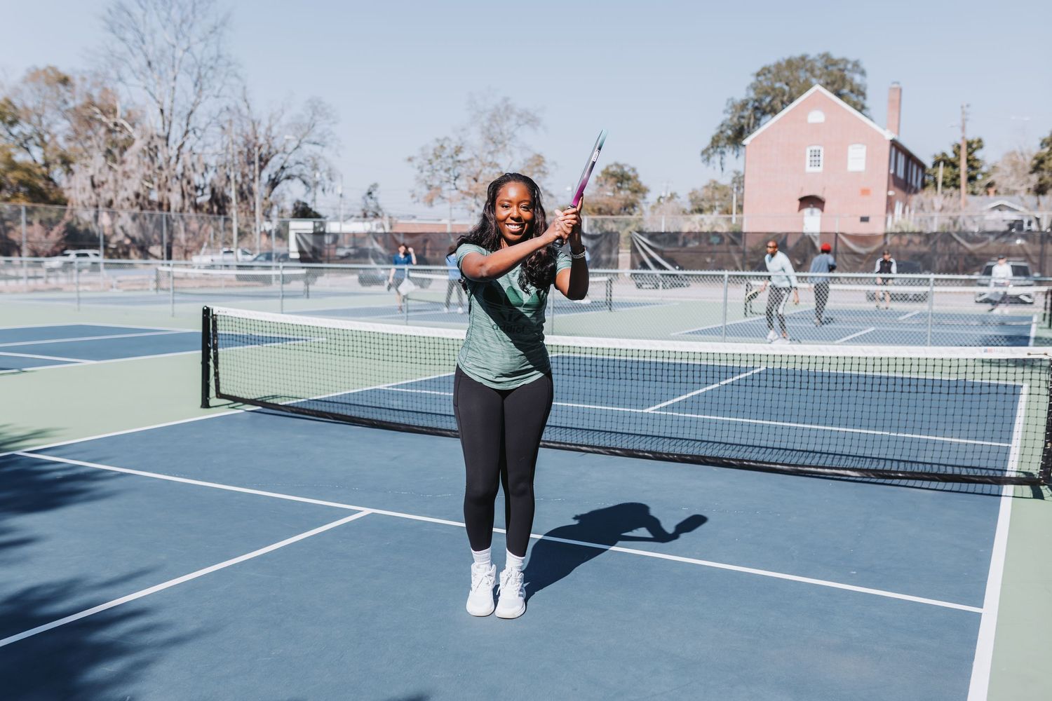 Woman smiling on blue pickleball court holding paddle overhead in sunny outdoor setting.