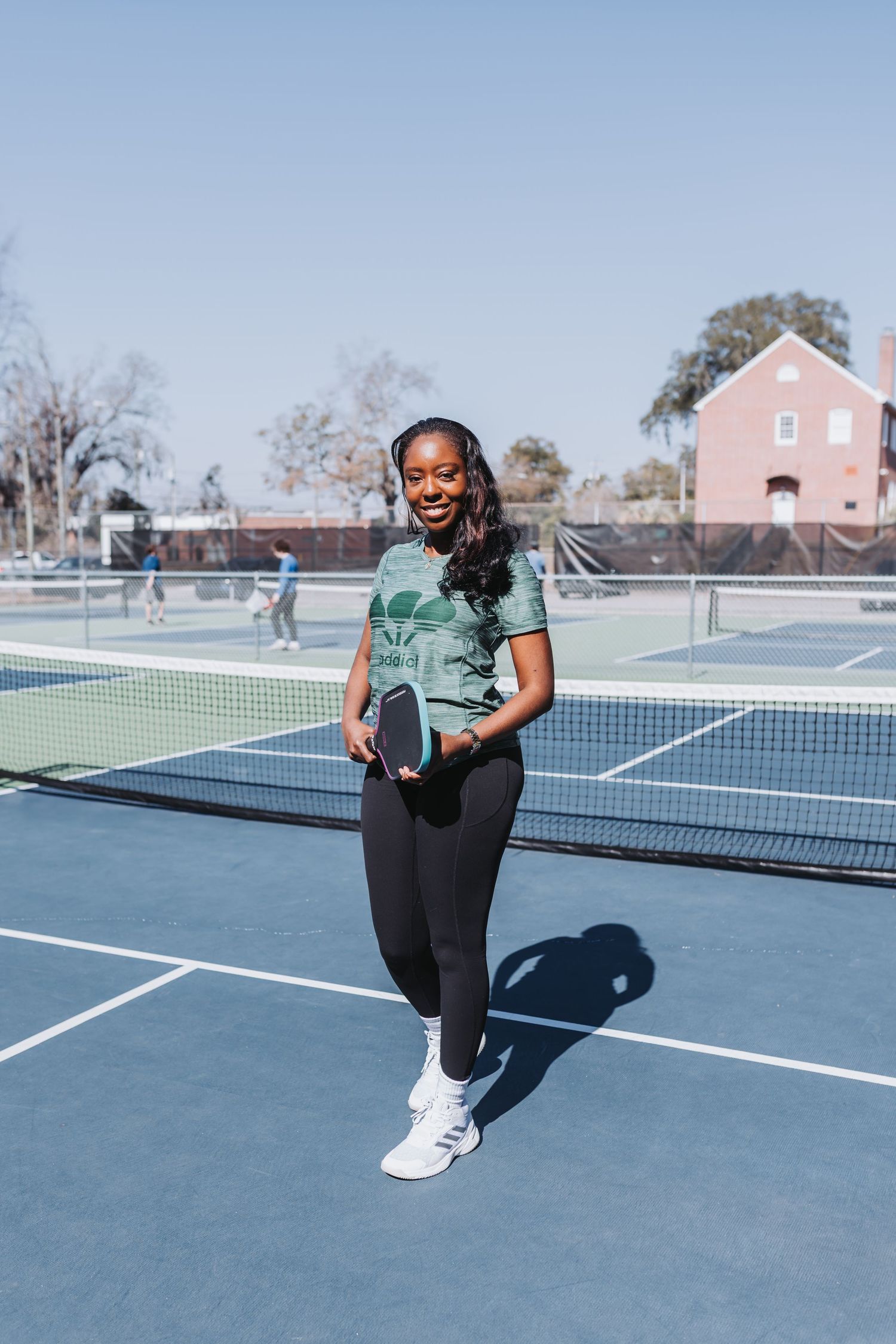 Smiling woman holding a pickleball paddle on a blue outdoor court, wearing headphones, green top, and black leggings.