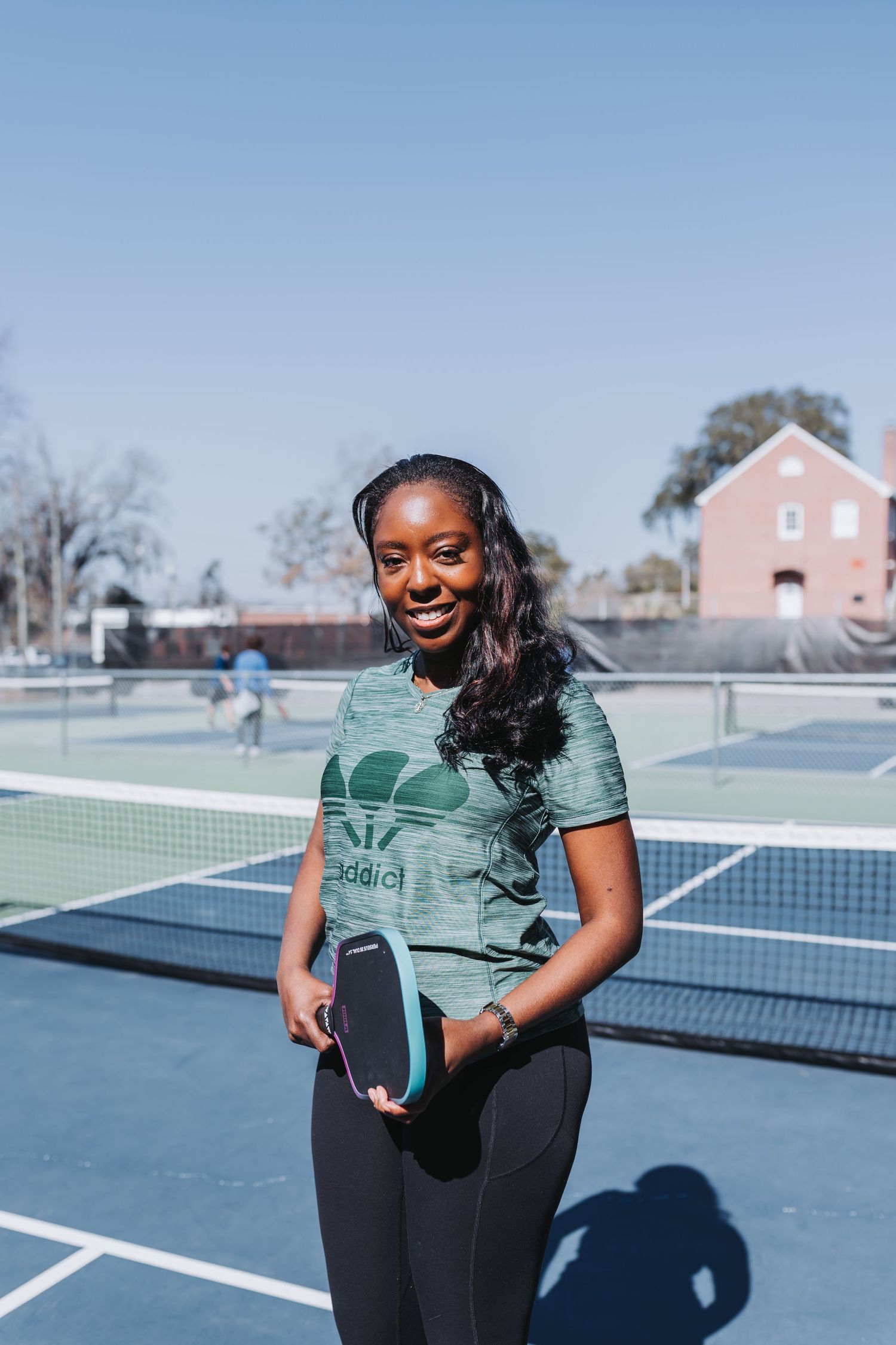Smiling woman holding a pickleball paddle on an outdoor court wearing an Adidas t-shirt and black leggings.