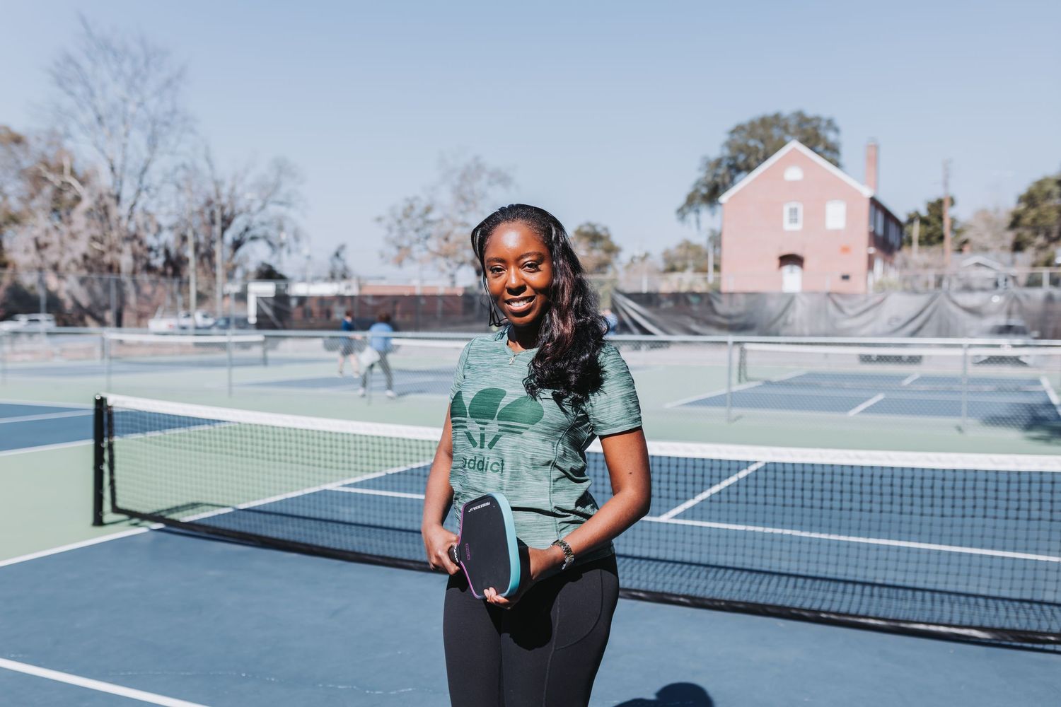 Woman smiling on blue pickleball court holding paddle, wearing green top and black leggings on sunny day.
