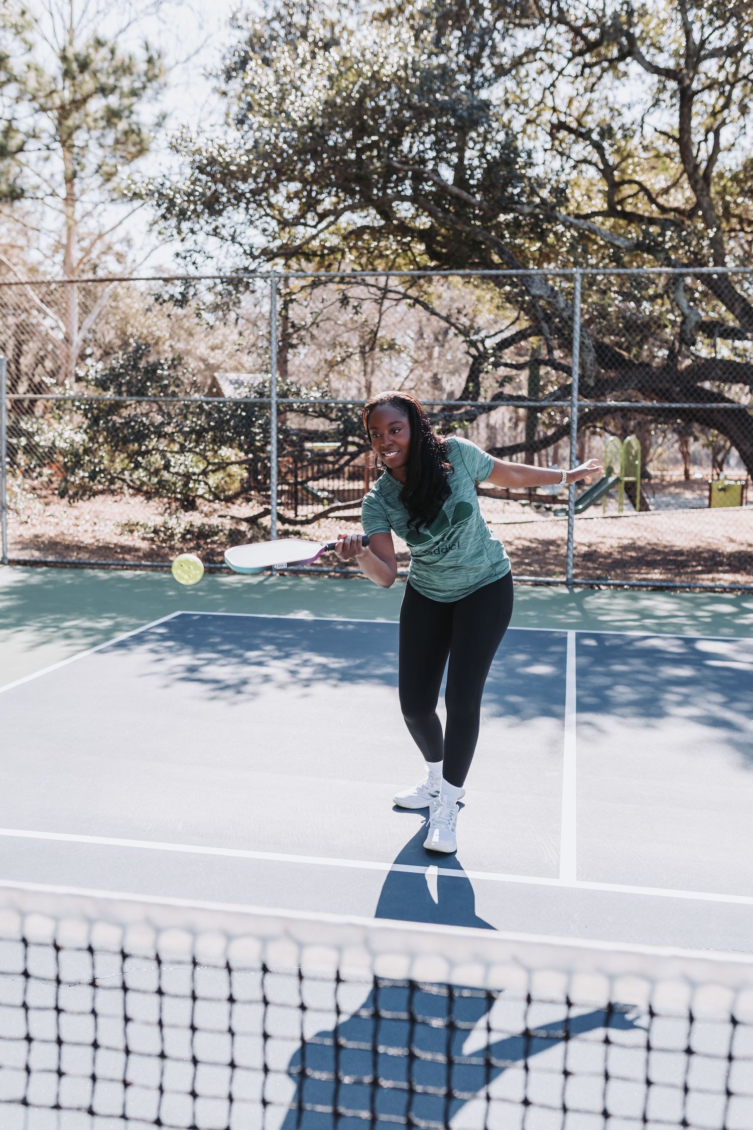 A woman in a green shirt and black leggings plays pickleball on an outdoor court on a sunny day.