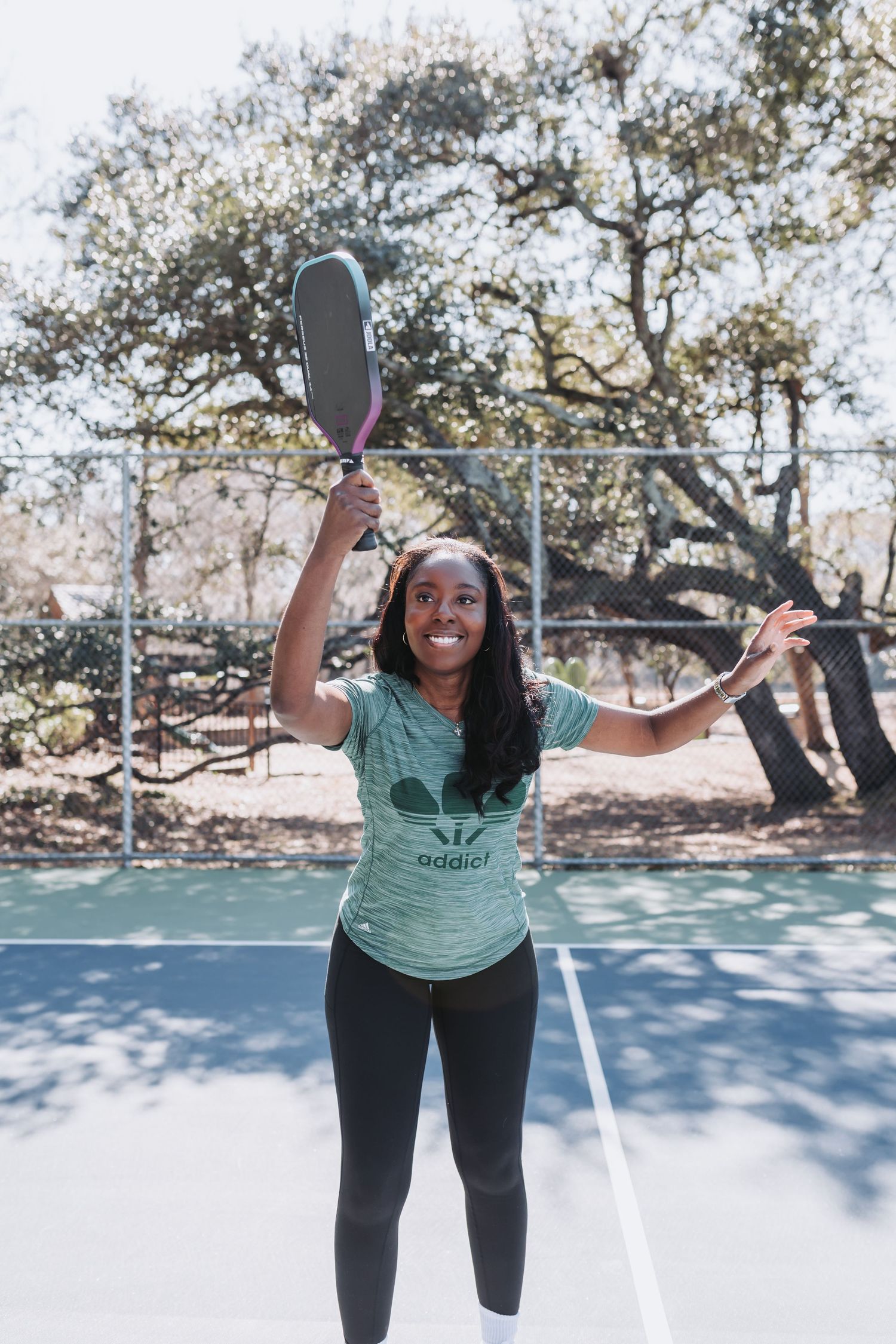 Woman smiling on pickleball court holding paddle overhead wearing green Adidas t-shirt and black leggings outdoors.