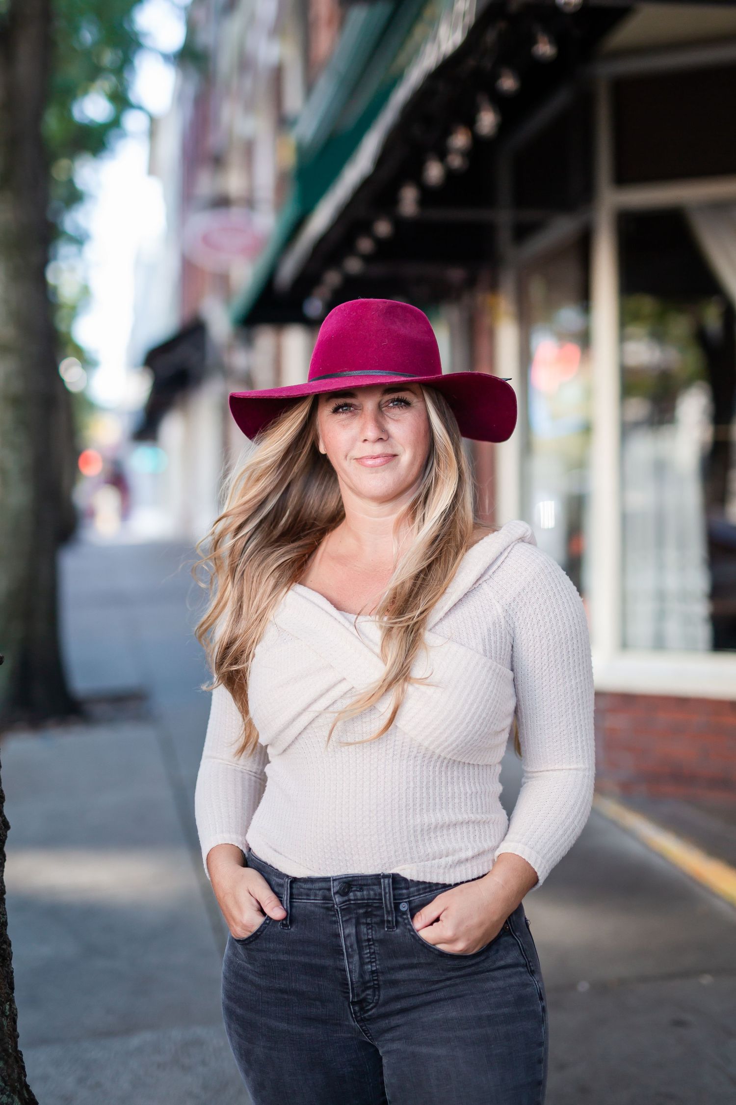 Woman in burgundy wide-brim hat and white ribbed top poses on urban sidewalk with hands in jeans pockets.
