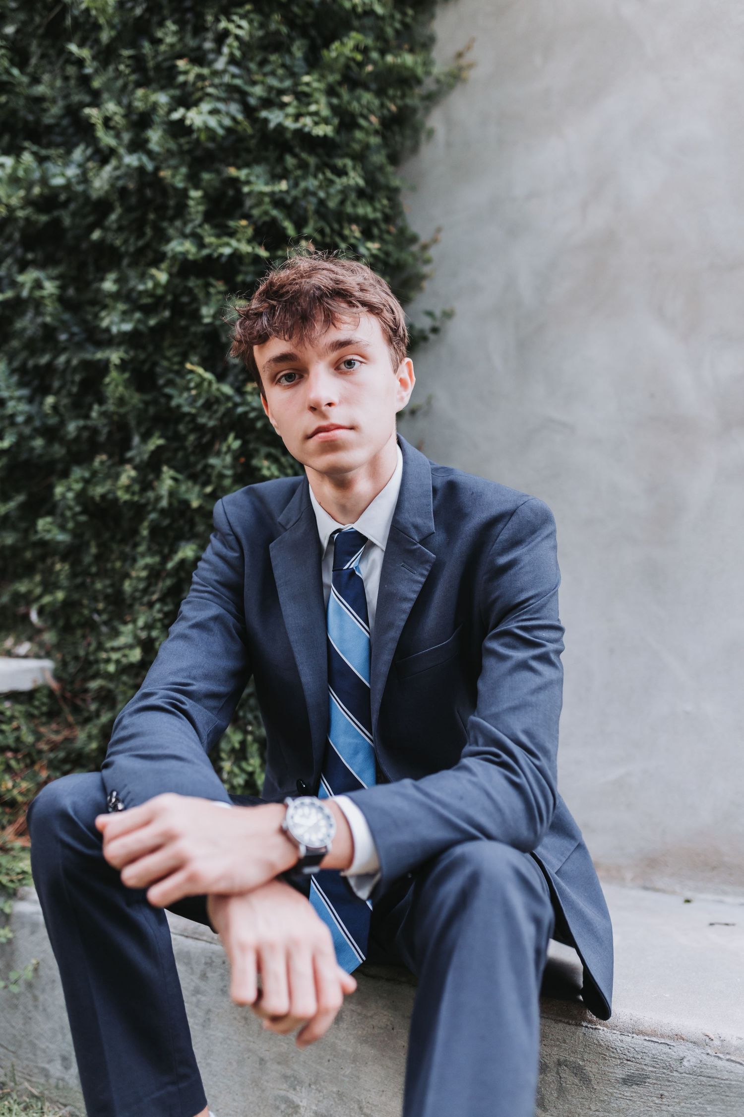 Young man in navy suit and striped tie sits on stone ledge outdoors, wearing a watch, looking at camera.