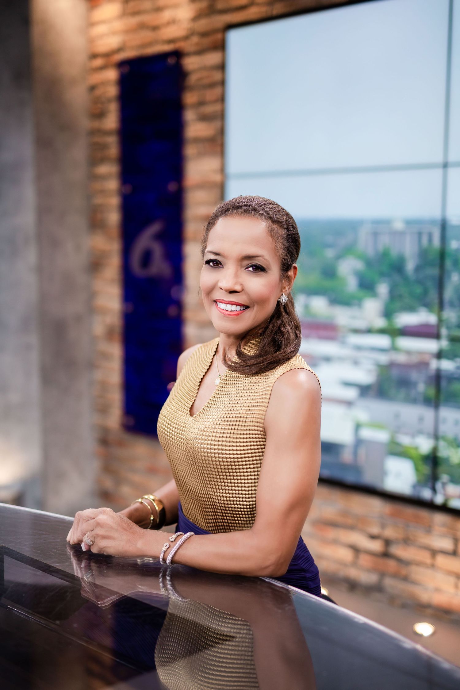 A woman in a gold sleeveless top smiles while leaning on a news desk in a modern TV studio.