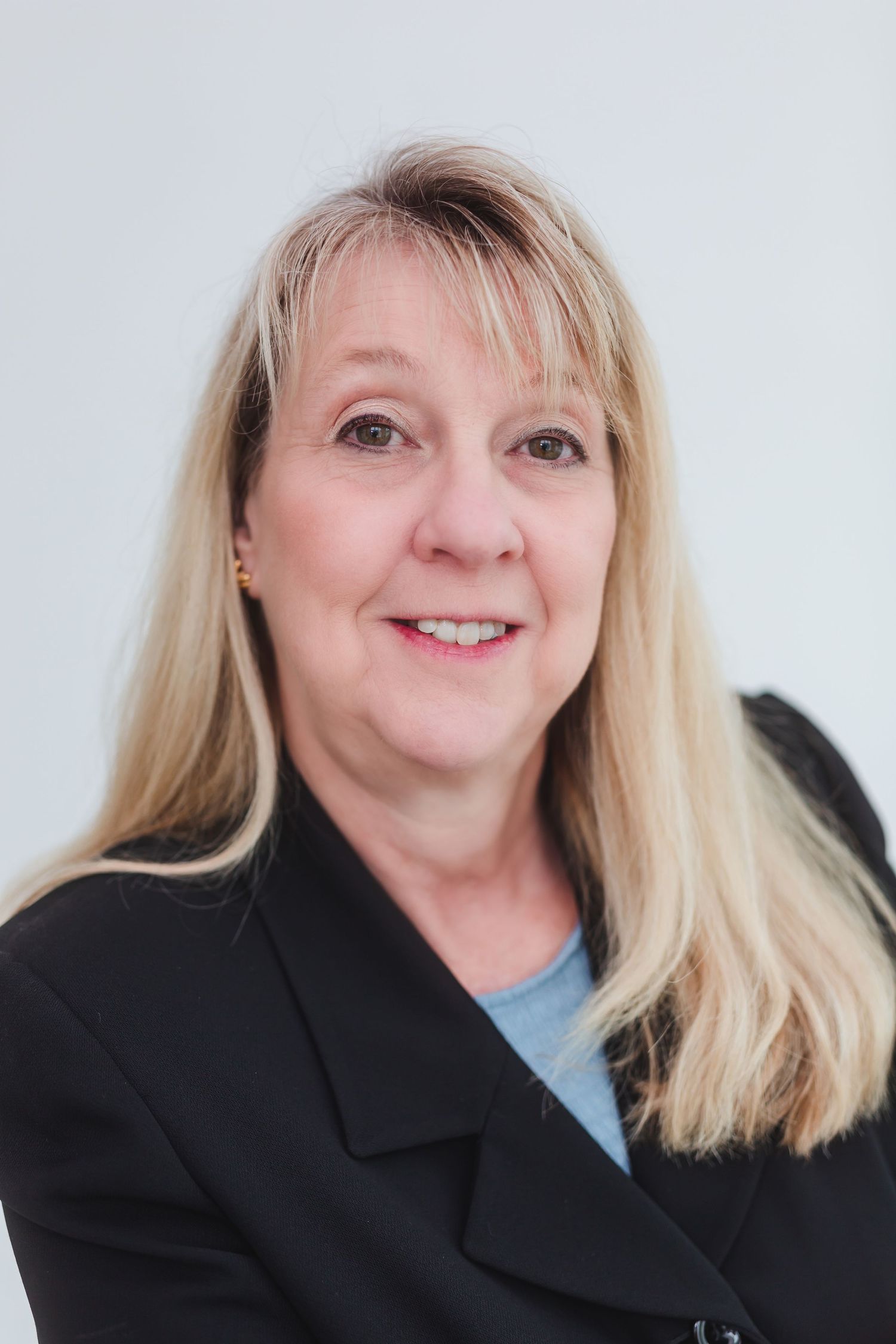 Professional headshot of a smiling middle-aged blonde woman wearing a black blazer against a light gray background.