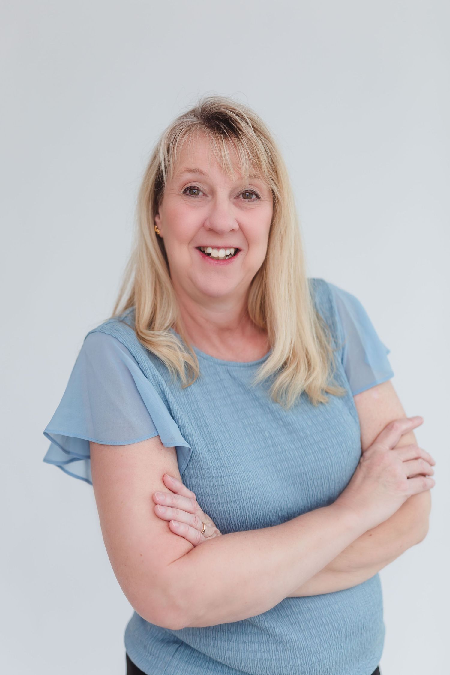 Smiling middle-aged blonde woman in blue top with arms crossed poses confidently against a light gray background.