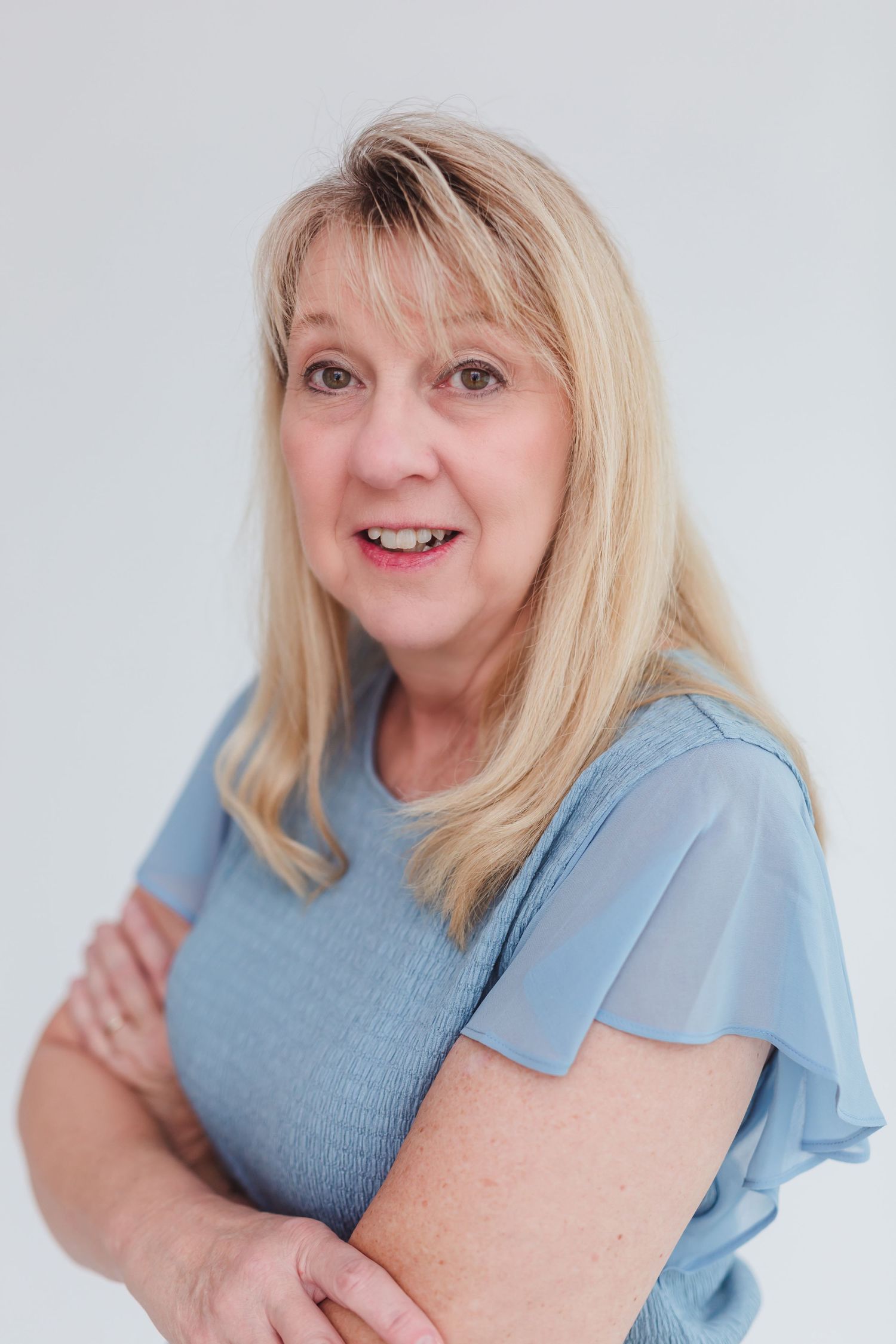 Smiling middle-aged blonde woman in a blue top with arms crossed against a white background.