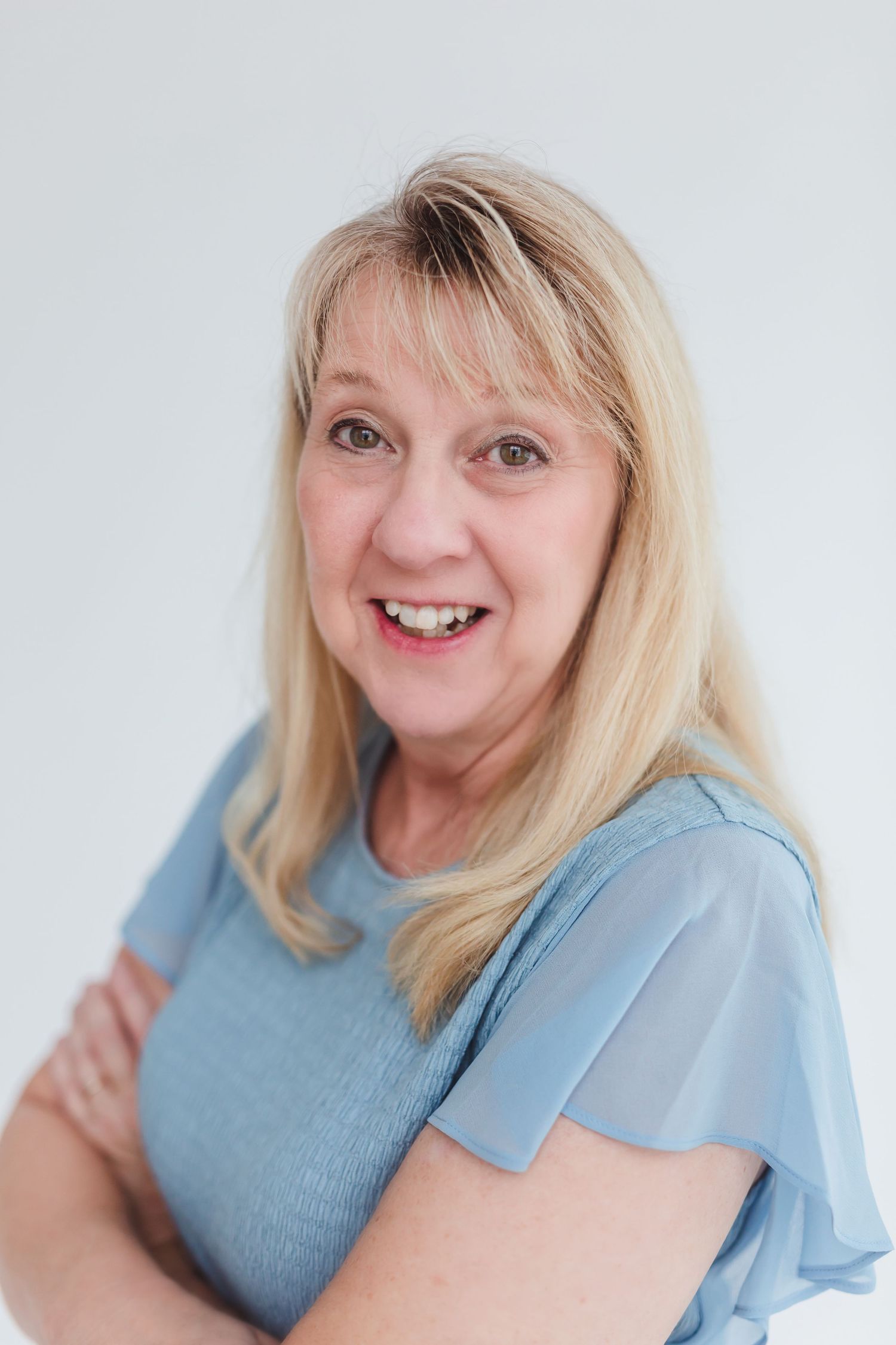 Smiling middle-aged blonde woman in a blue top with arms crossed against a white background.