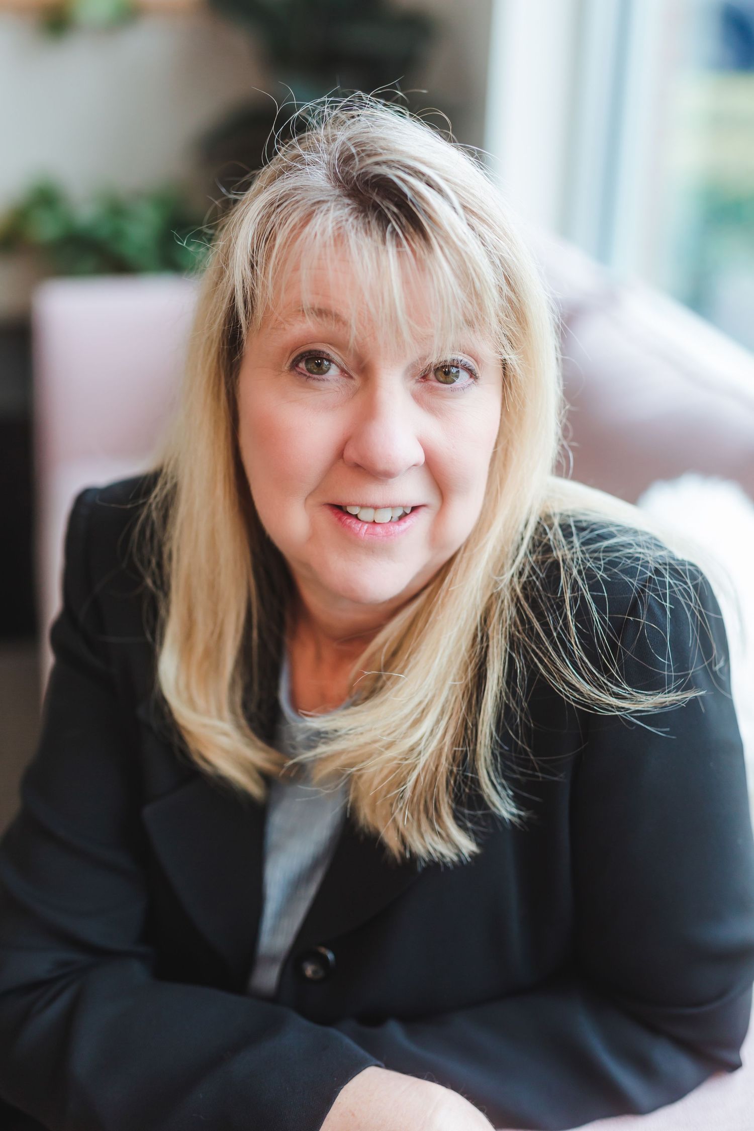 Professional headshot of a smiling blonde woman in a black blazer, seated in a bright modern interior setting.