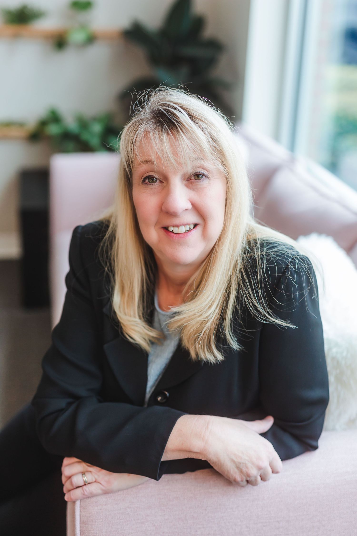 Smiling blonde woman in black blazer poses on pink sofa in bright modern interior setting.