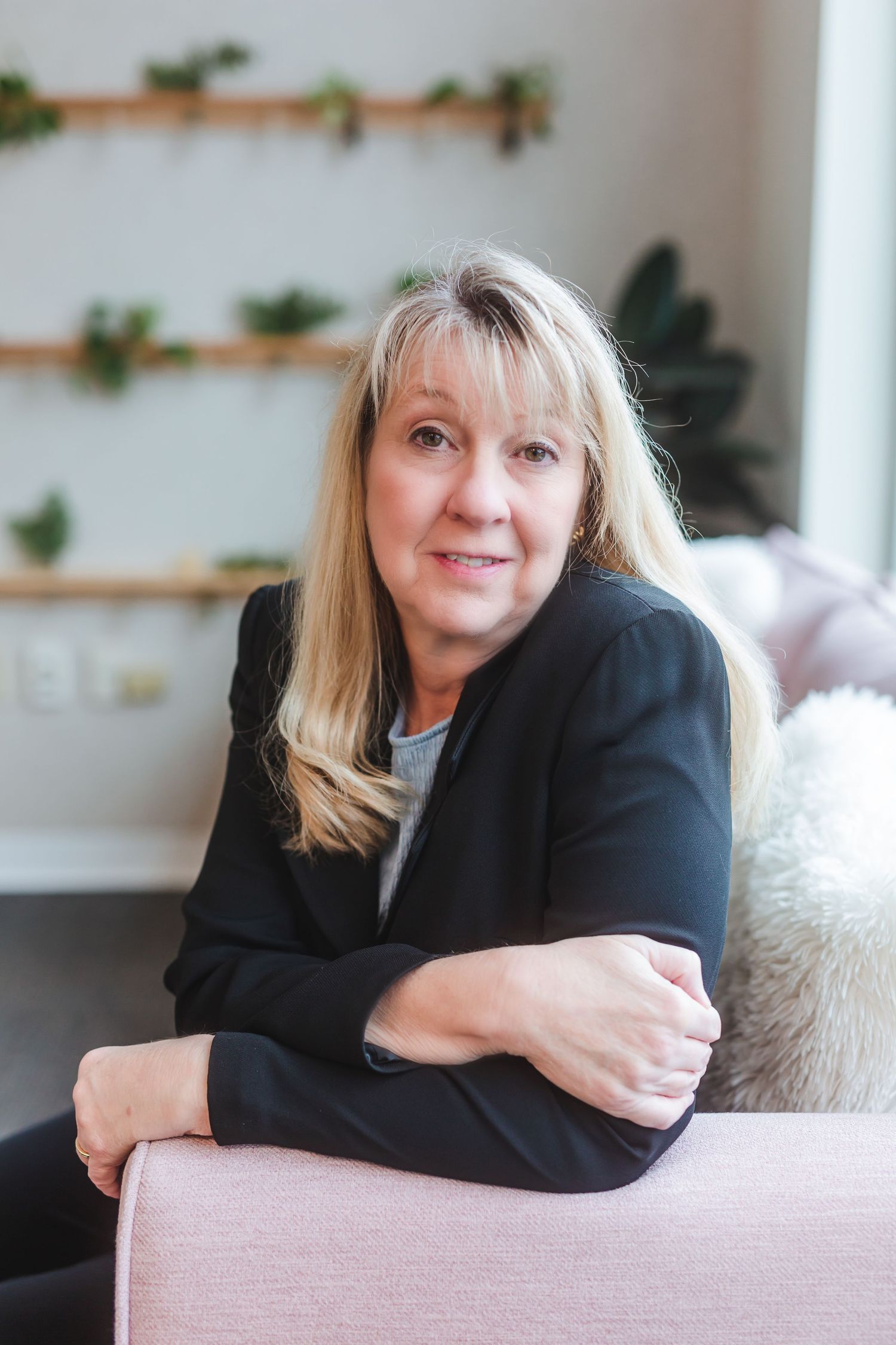 Middle-aged blonde woman in black blazer sitting relaxed on pink sofa in modern interior with plant wall decor.