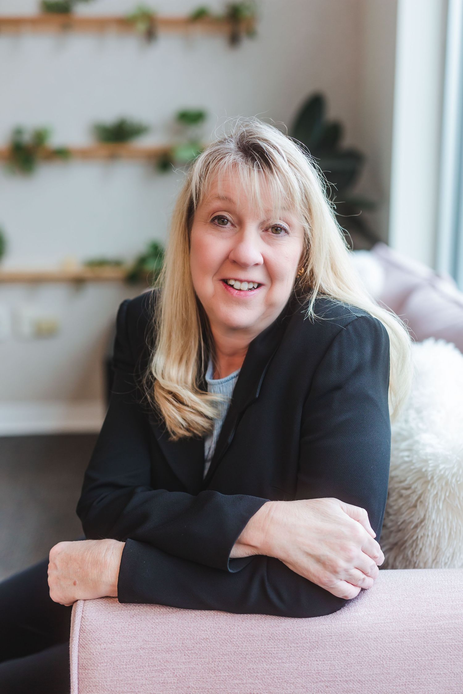 Smiling blonde woman in black blazer relaxing on pink sofa in modern interior with wall plants.