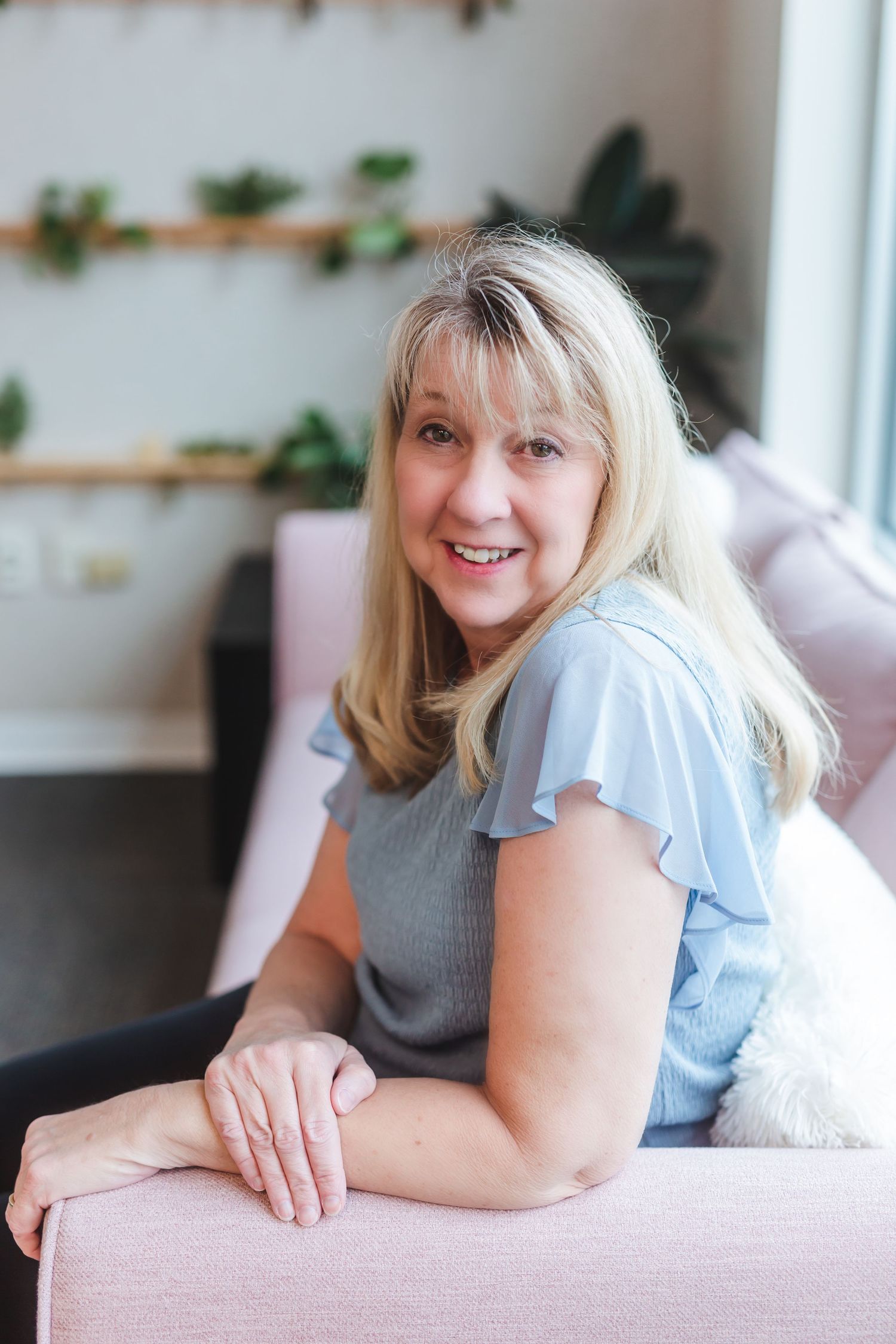 Smiling middle-aged blonde woman in blue top sitting on pink sofa in bright modern room with plants.