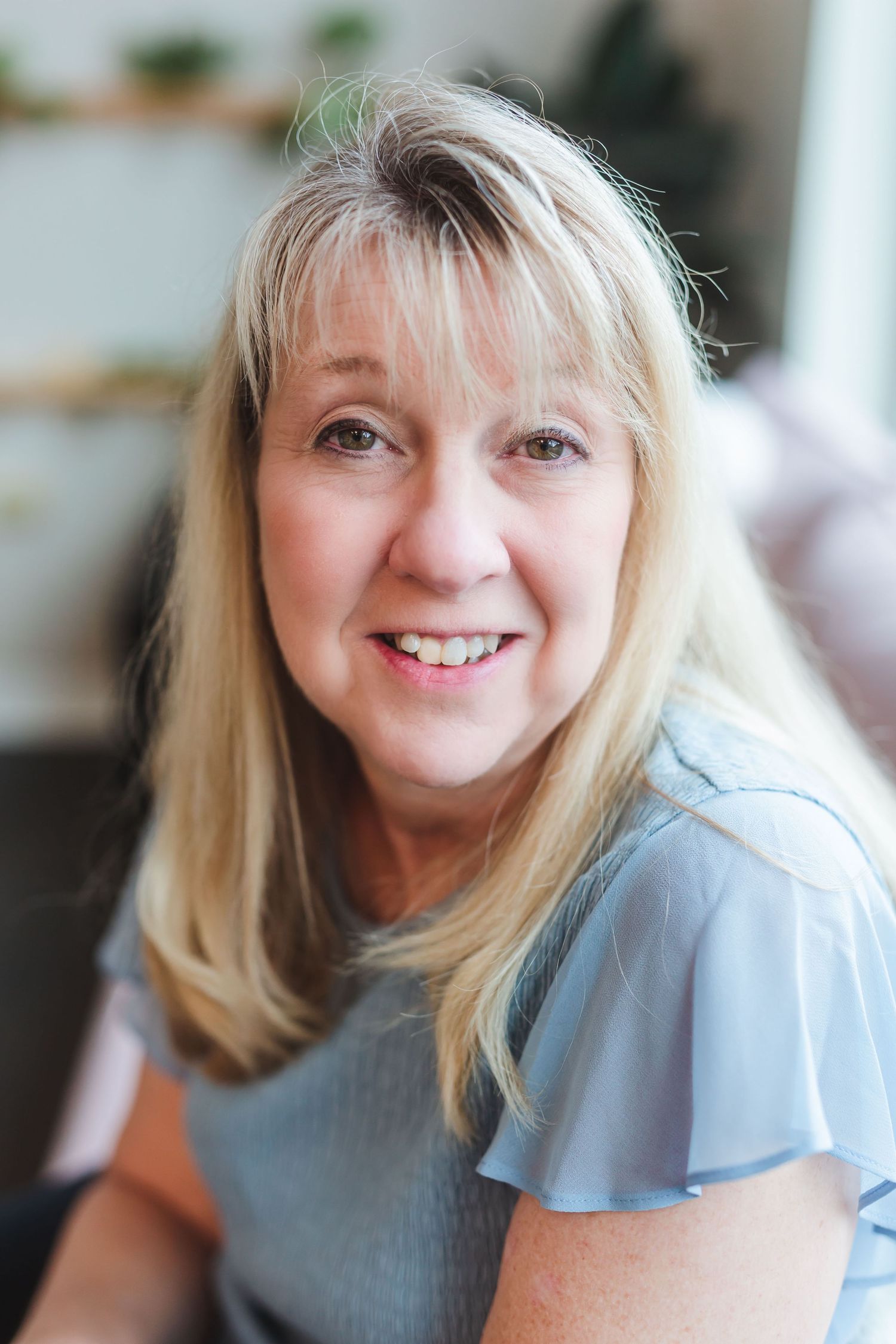 Smiling middle-aged blonde woman in a light blue top poses for a portrait in a bright, softly blurred indoor setting.
