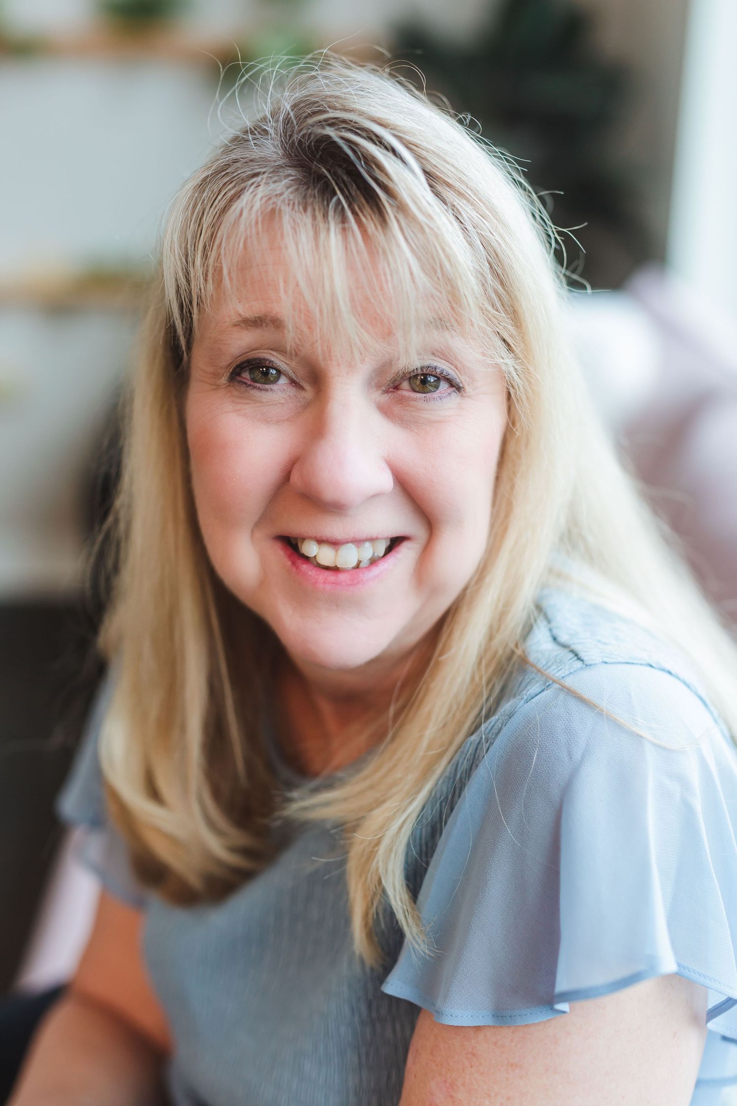 Smiling middle-aged blonde woman in a light blue top poses for a portrait in a bright, softly blurred indoor setting.