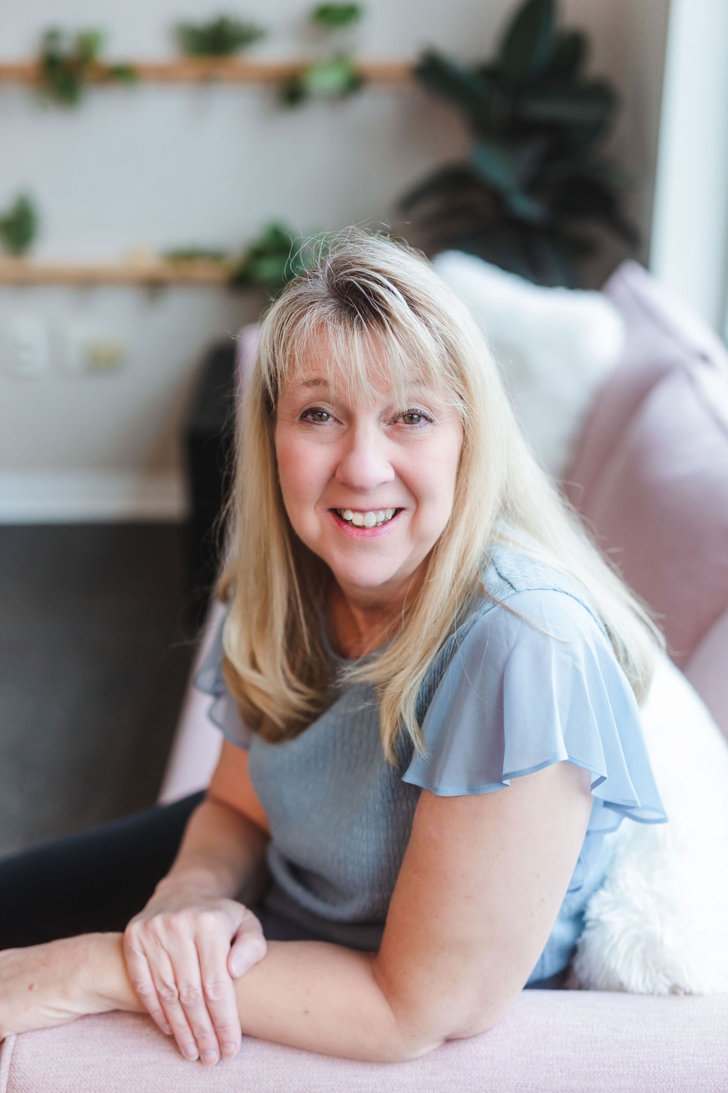 Smiling middle-aged blonde woman in blue top relaxing on pink sofa with green plants on shelves behind her.