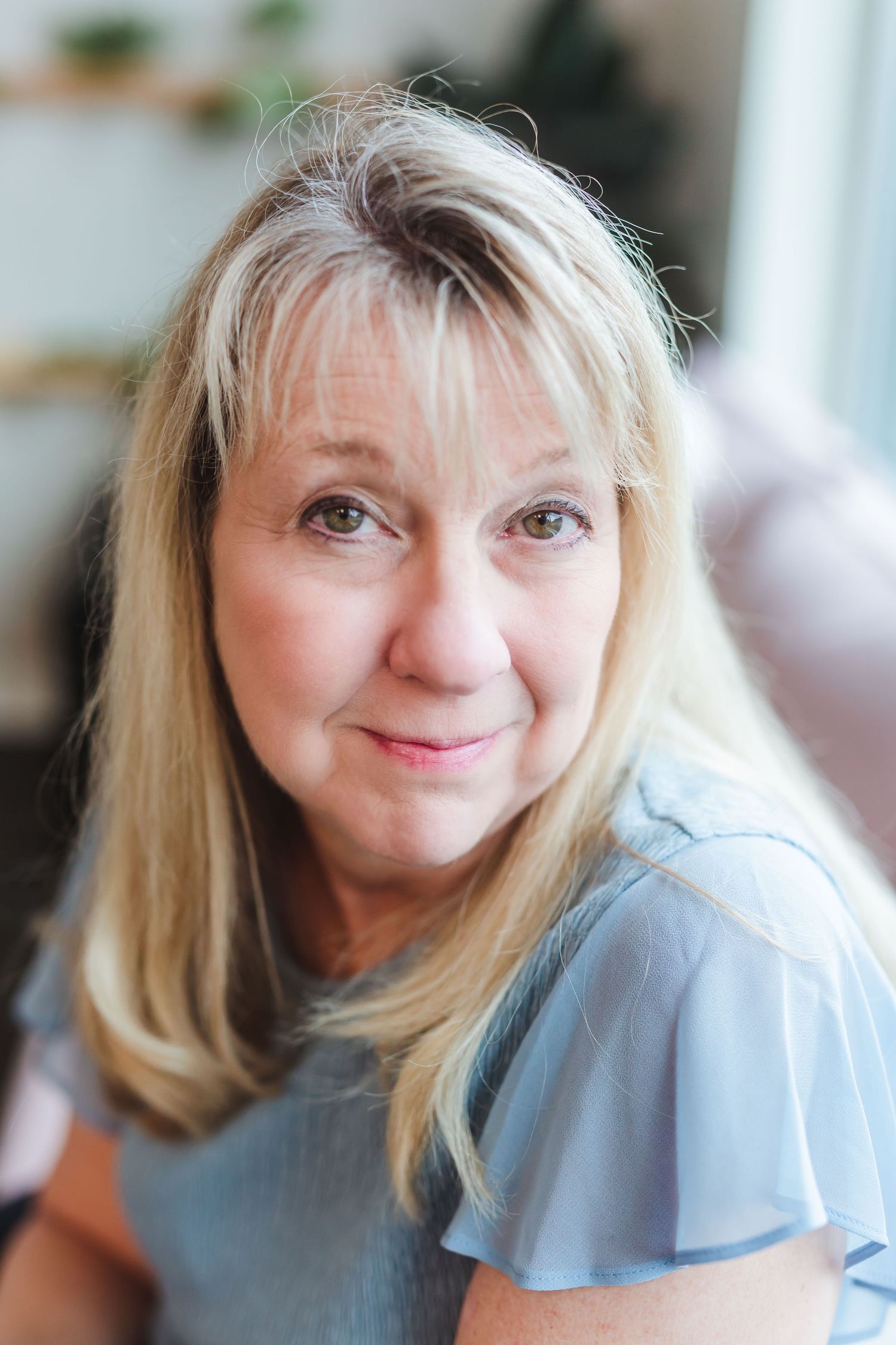 Middle-aged blonde woman in light blue top smiling warmly indoors with soft natural lighting.