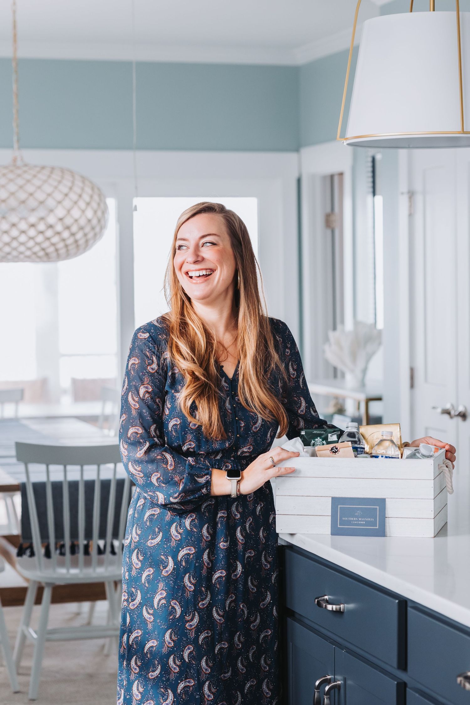 Smiling woman in blue floral dress holding a gift box in a modern kitchen with navy cabinets and pendant lights.